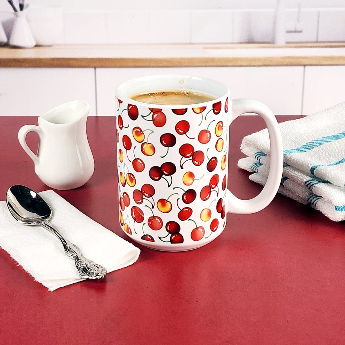 15oz Cherries Mug from Amy's Coffee Mugs on a red counter in a kitchen next to a spoon, creamer pitcher, and a towel.