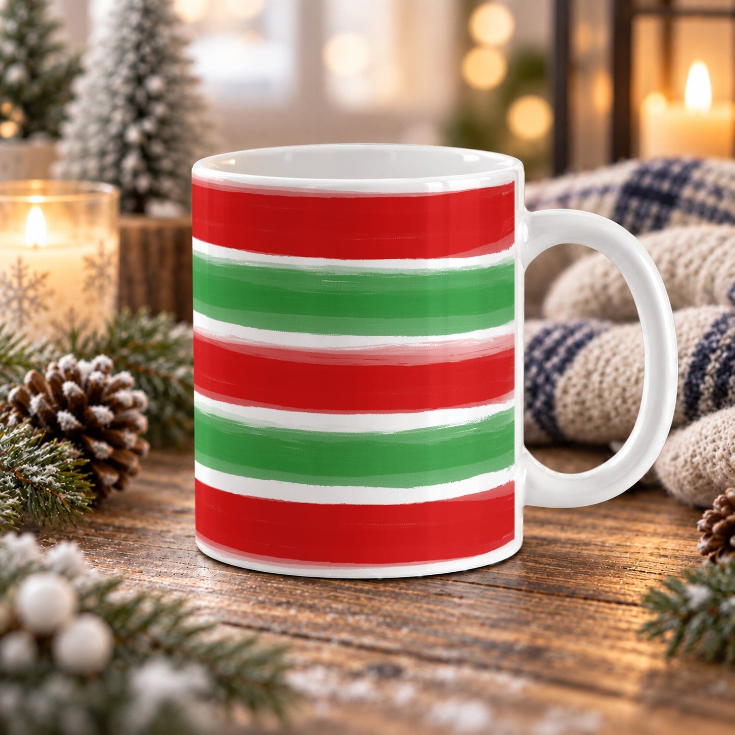 11 oz Christmas mug with red and green painted brushstroke stripes wrapping around the white ceramic mug, on a wooden table with winter decor around it including pine cones, pine boughs, candles, and a cozy plaid blanket.