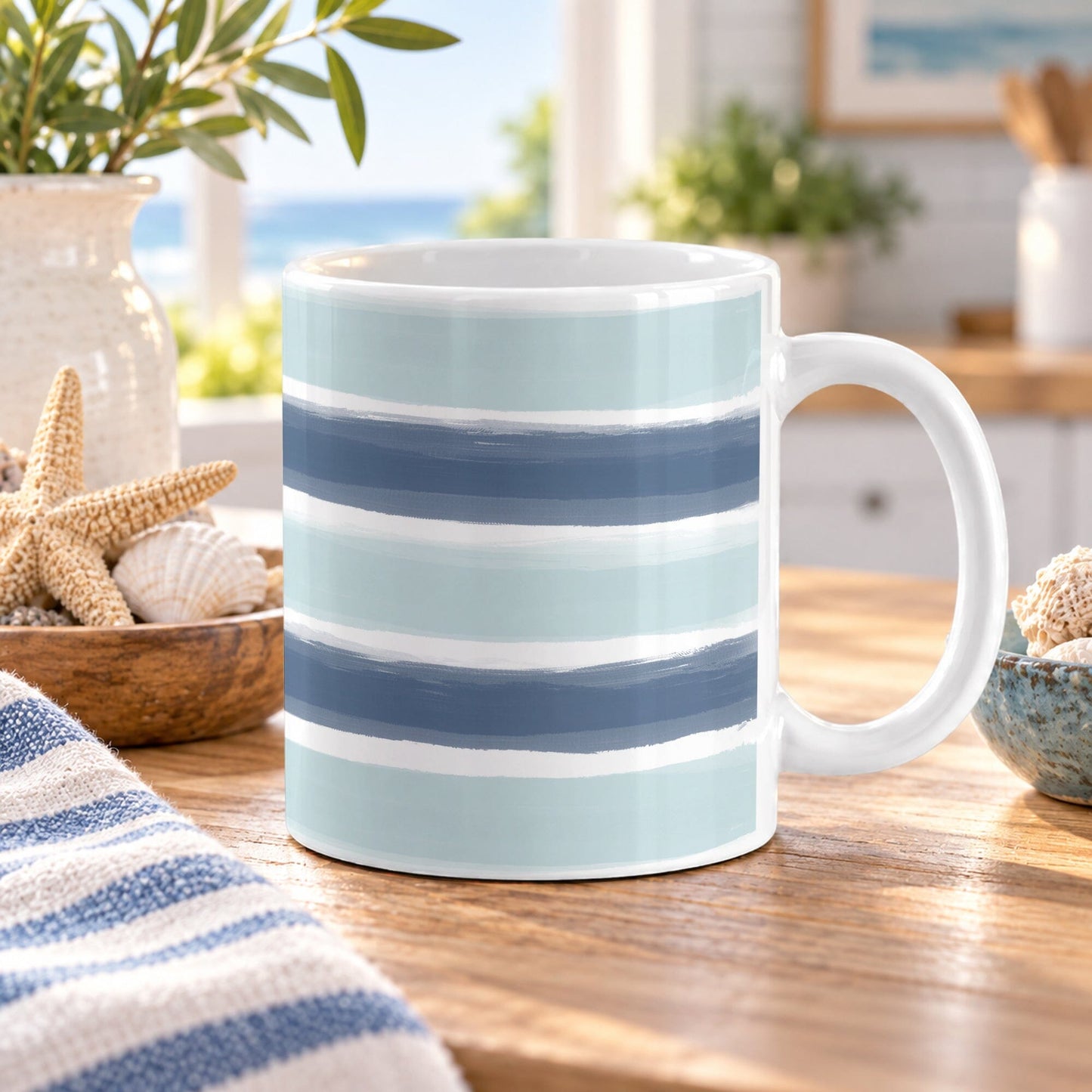 11 oz coastal blue coffee mug with horizontal navy and light blue brushstroke stripes over the glossy white ceramic mug, styled on a wooden table with a striped towel and seashells in a bright, coastal-themed kitchen setting.