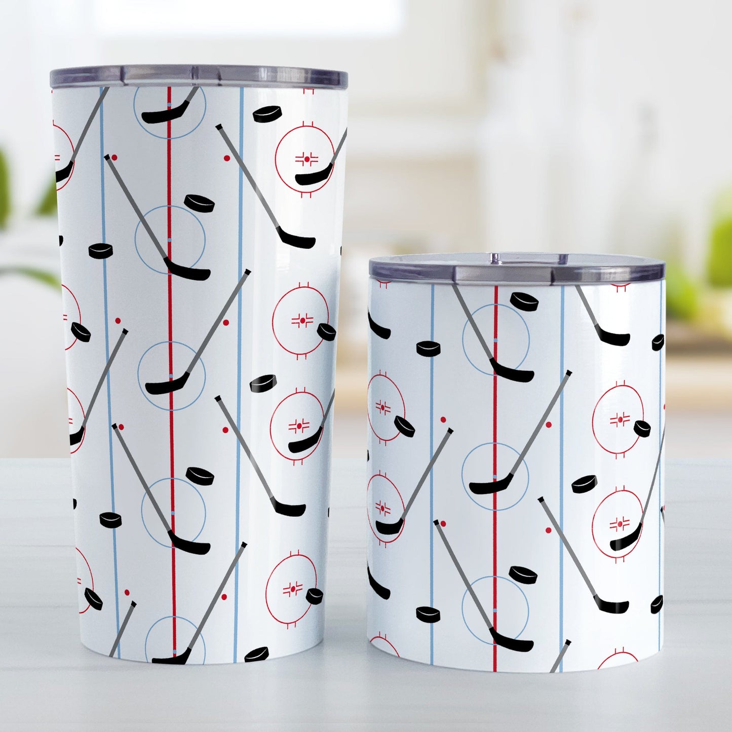 Hockey Rink Pattern Tumbler Cup (20oz and 10oz) at Amy's Coffee Mugs. A stainless steel tumbler cup designed with hockey rink red and blue lines, coupled with hockey sticks and hockey pucks, a pattern that wraps around the cup. Photo shows both sized cups on a table next to each other.