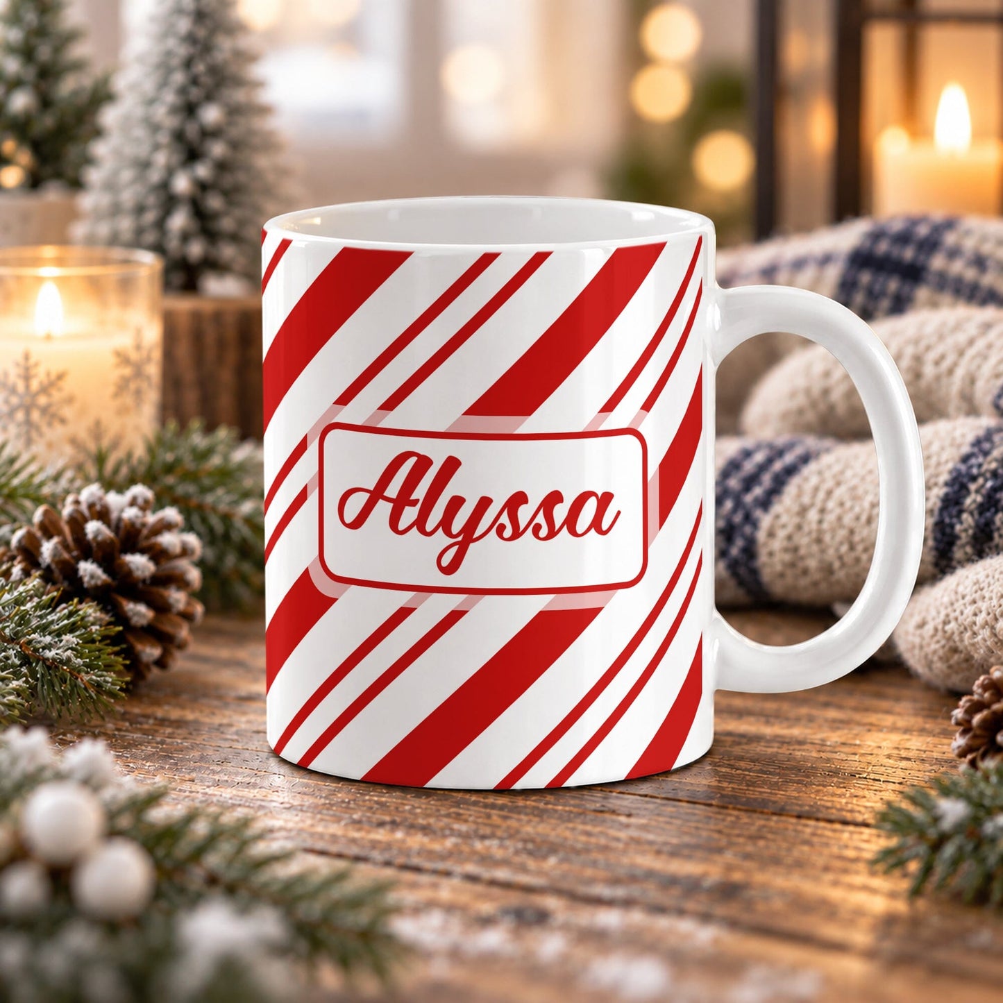 11 oz personalized candy cane stripe mug with red and white diagonal stripes and custom name in red script on both sides, on a wooden table with winter decor around it including pine cones, pine boughs, candles, and a cozy plaid blanket.