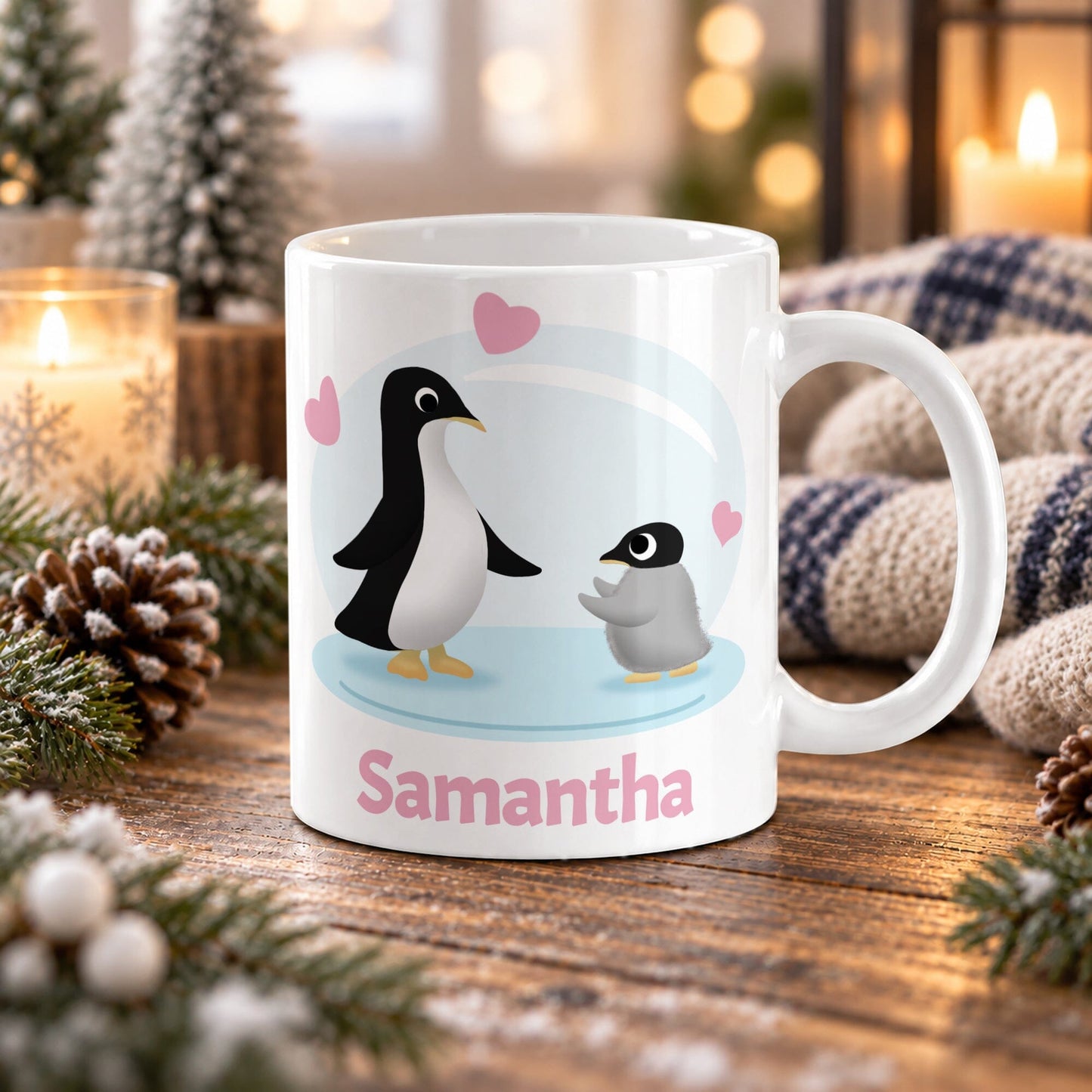11 oz personalized penguin mug with adult penguin, fluffy baby chick, pink hearts, soft blue background, and custom name in pink, on a wooden table with winter decor around it including pine cones, pine boughs, candles, and a cozy plaid blanket.