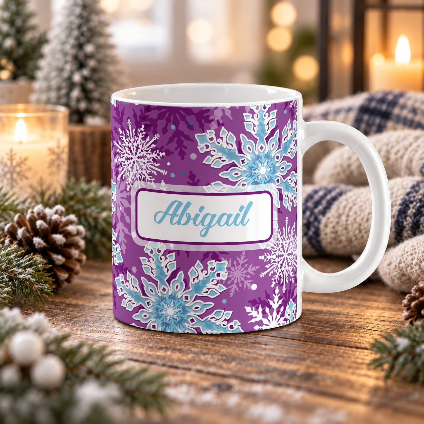 11 oz personalized purple snowflake mug with blue and white snowflakes, aqua blue custom name, and purple winter background, on a wooden table with winter decor around it including pine cones, pine boughs, candles, and a cozy plaid blanket.