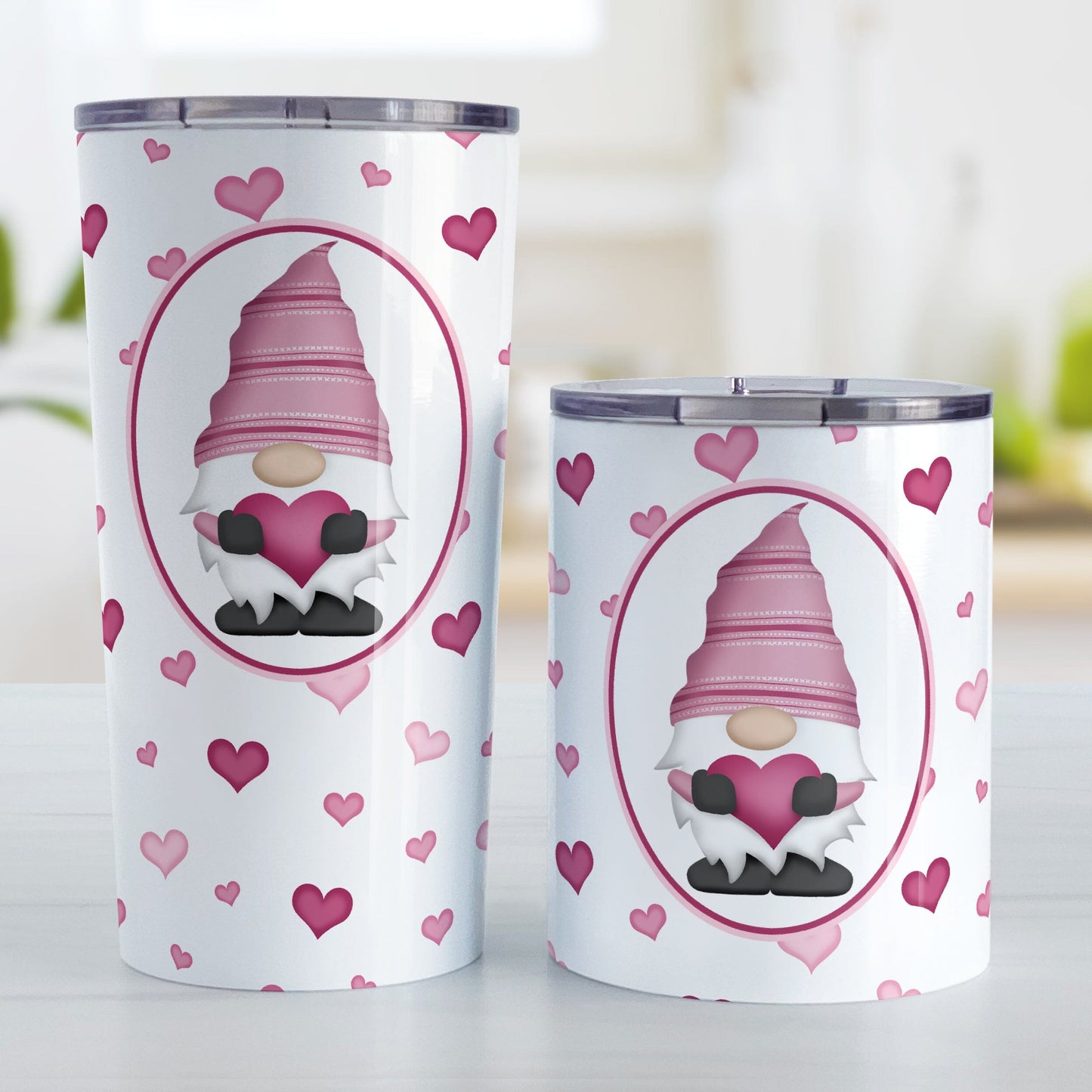 Pink Gnome Dainty Hearts Tumbler Cup (20oz or 10oz) at Amy's Coffee Mugs. Stainless steel tumbler cups designed with an adorable pink gnome holding a heart in a white oval over a pattern of cute and dainty hearts in different shades of pink that wrap around the cups. Photo shows both sized cups on a table next to each other.