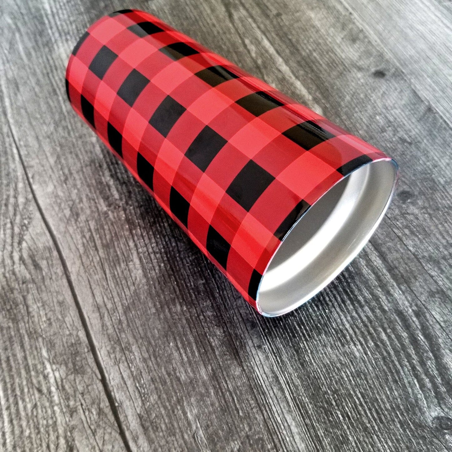 A 20oz tumbler cup designed with a red and black buffalo plaid (buffalo check) pattern that wraps around the cup. Photo shows the cup laying on its side on a weathered wood surface.