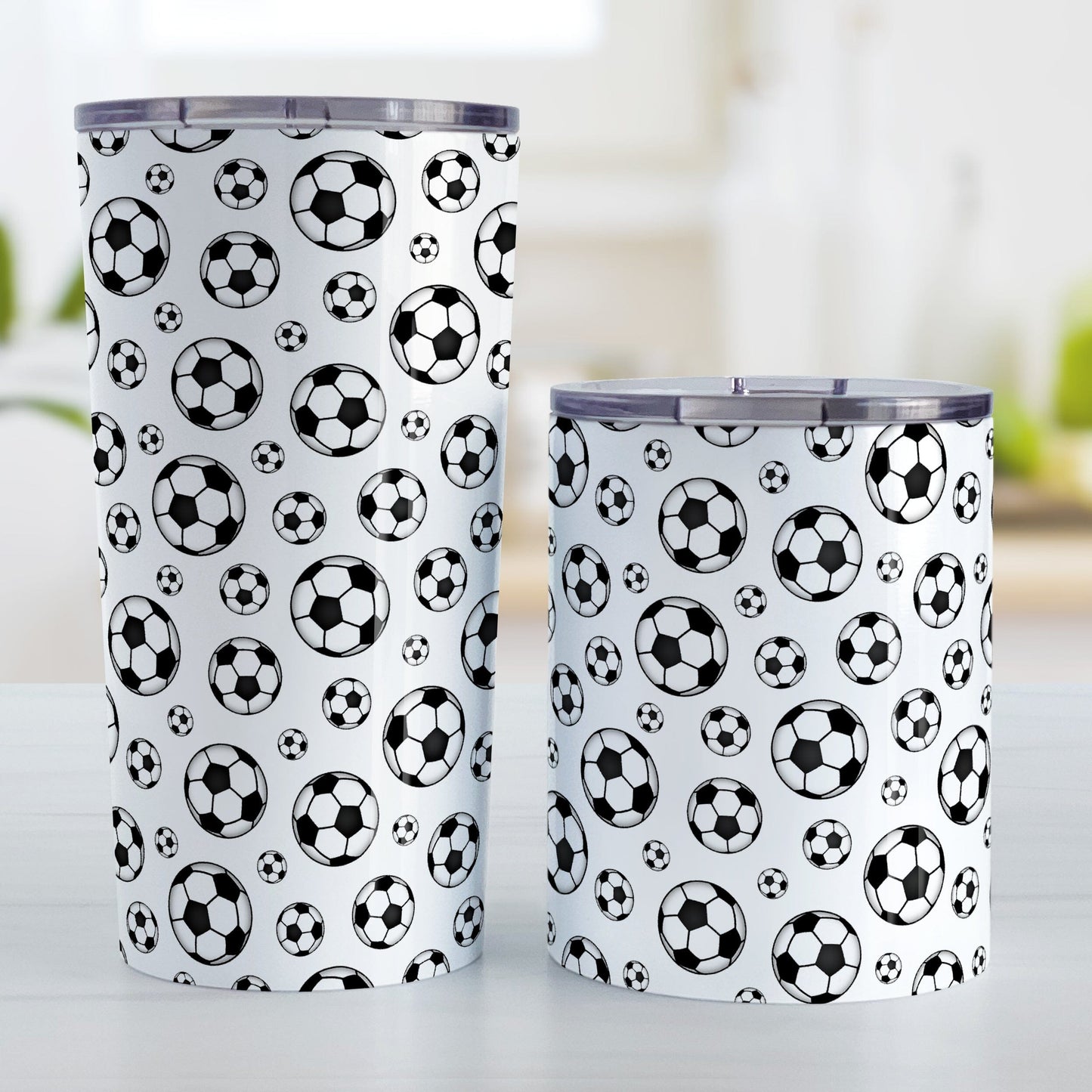 Soccer Balls Pattern Tumbler Cup (20oz and 10oz) at Amy's Coffee Mugs. A stainless steel tumbler cup designed with black and white soccer balls in varying sizes in a pattern that wraps around the cup. Photo shows both sized mugs on a table next to each other.