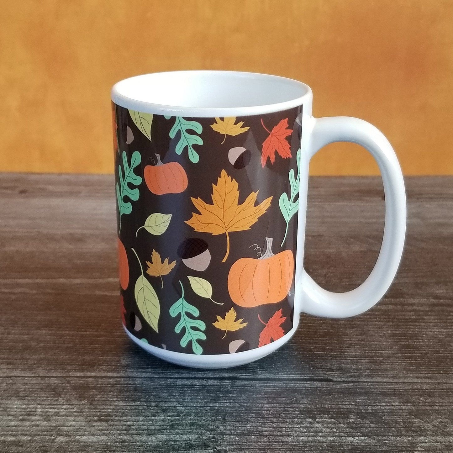Autumn Pumpkins and Leaves Pattern - Fall Mug (15oz) on orange and weathered wood background. Amy's Coffee Mugs. A ceramic coffee mug designed with a pattern of pumpkins, acorns, and leaves in orange, yellow, and green over a brown background color that wraps around the mug up to the handle.