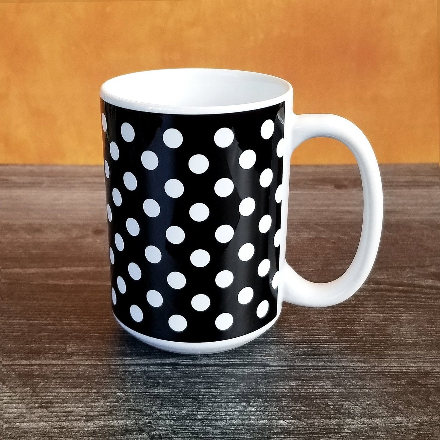 Black Polka Dot Mug (15oz, on orange and weathered wood background) at Amy's Coffee Mugs. A ceramic coffee mug designed with a pattern of white polka dots over a black background color that wraps around the mug up to the handle.