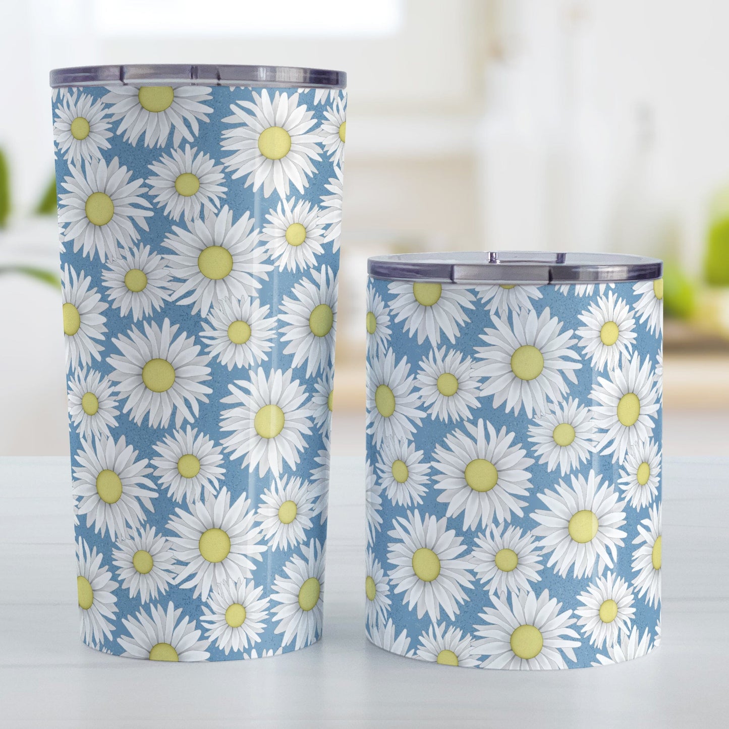 Blue Daisy Pattern Tumbler Cups (20oz or 10oz) at Amy's Coffee Mugs. Stainless steel tumbler cups designed with a pretty pattern of white daisy flowers with yellow centers over a speckled blue background that wraps around the cups. Photo shows both sized cups on a table next to each other.