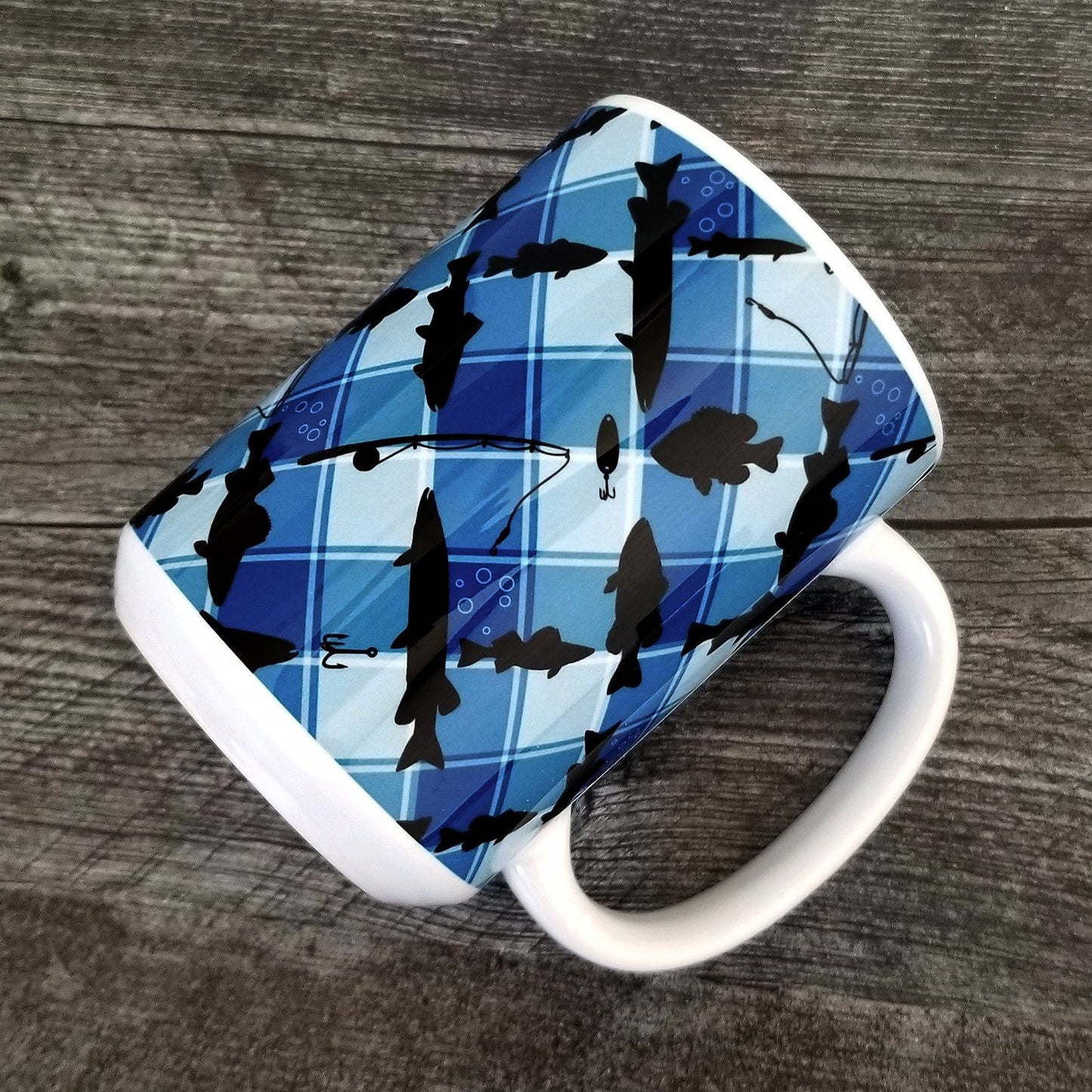 Blue Fishing Plaid Pattern Mug at Amy's Coffee Mugs. A ceramic coffee mug designed with black silhouettes of fish, fishing poles, and lures diagonally arranged over a blue plaid pattern that wraps around the mug up to the handle. Photo shows the mug laying on a wood surface.