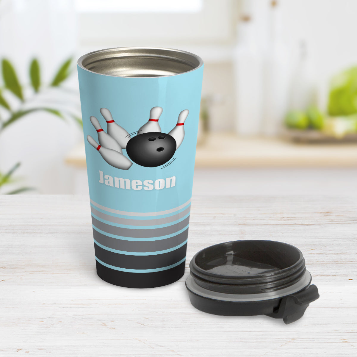 Bowling Ball and Pins Blue - Personalized Bowling Travel Mug (15oz) at Amy's Coffee Mugs. A travel mug designed with an illustration of a bowling ball striking bowling pins over a light blue background color that wraps around the travel mug. Along the bottom of the mug are gray stripes in varying shades creating a dynamic look. Your custom name is printed in white below the bowling ball and pins. Photo shows the mug open on a table with its lid beside it.