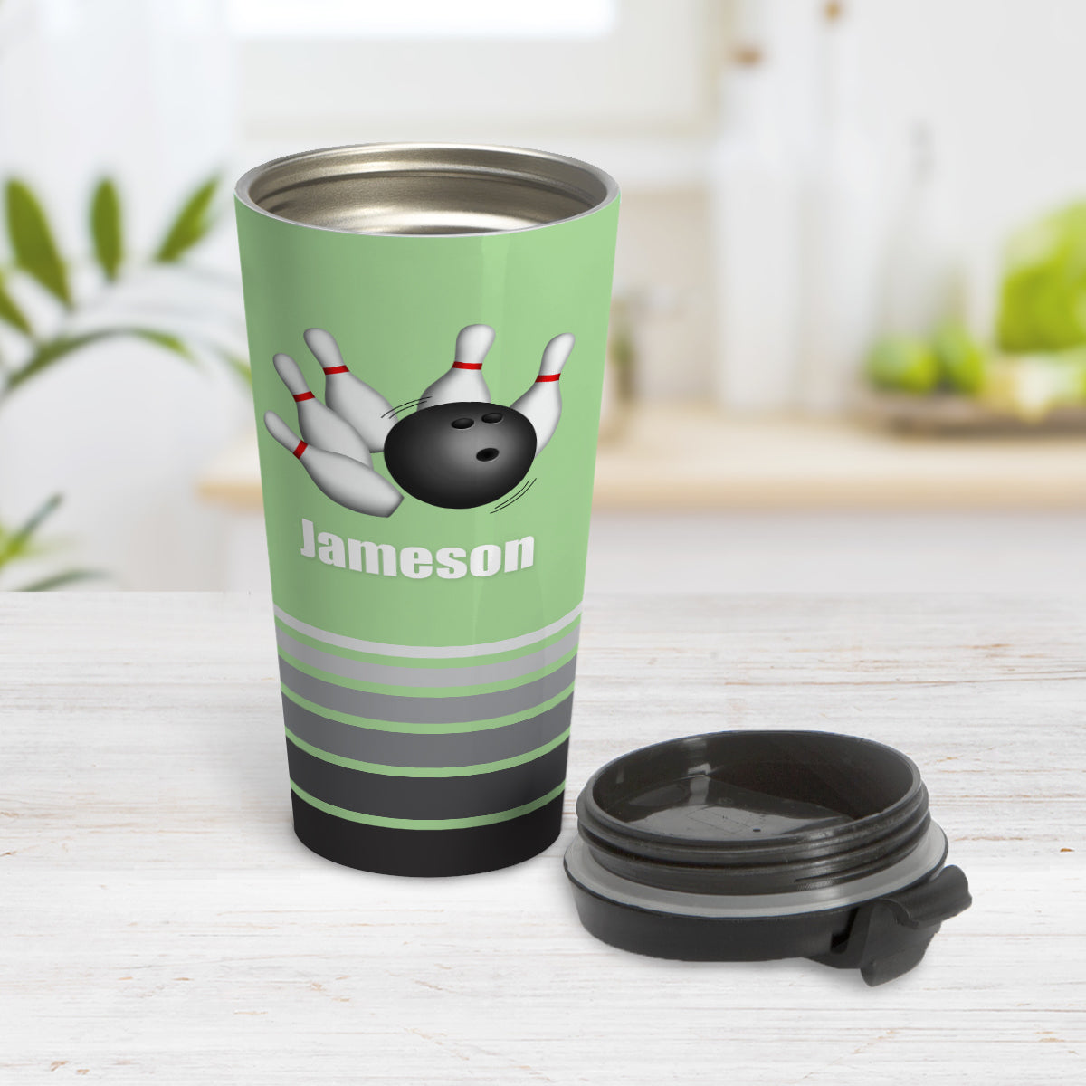 Bowling Ball and Pins Green - Personalized Bowling Travel Mug (15oz) at Amy's Coffee Mugs. A travel mug designed with an illustration of a bowling ball striking bowling pins over a green background color that wraps around the travel mug. Along the bottom of the mug are gray stripes in varying shades creating a dynamic look. Your custom name is printed in white below the bowling ball and pins. Photo shows the mug open on a table with its lid beside it.