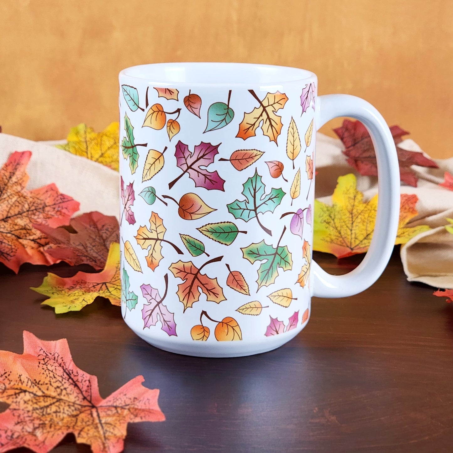 Changing Leaves Fall Mug (15oz) on a cherry wood table with leaves and a beige linen towel in front of an orange wall.