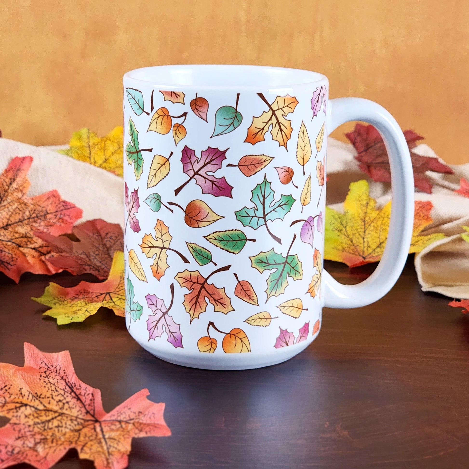 Changing Leaves Fall Mug (15oz) on a cherry wood table with leaves and a beige linen towel in front of an orange wall.