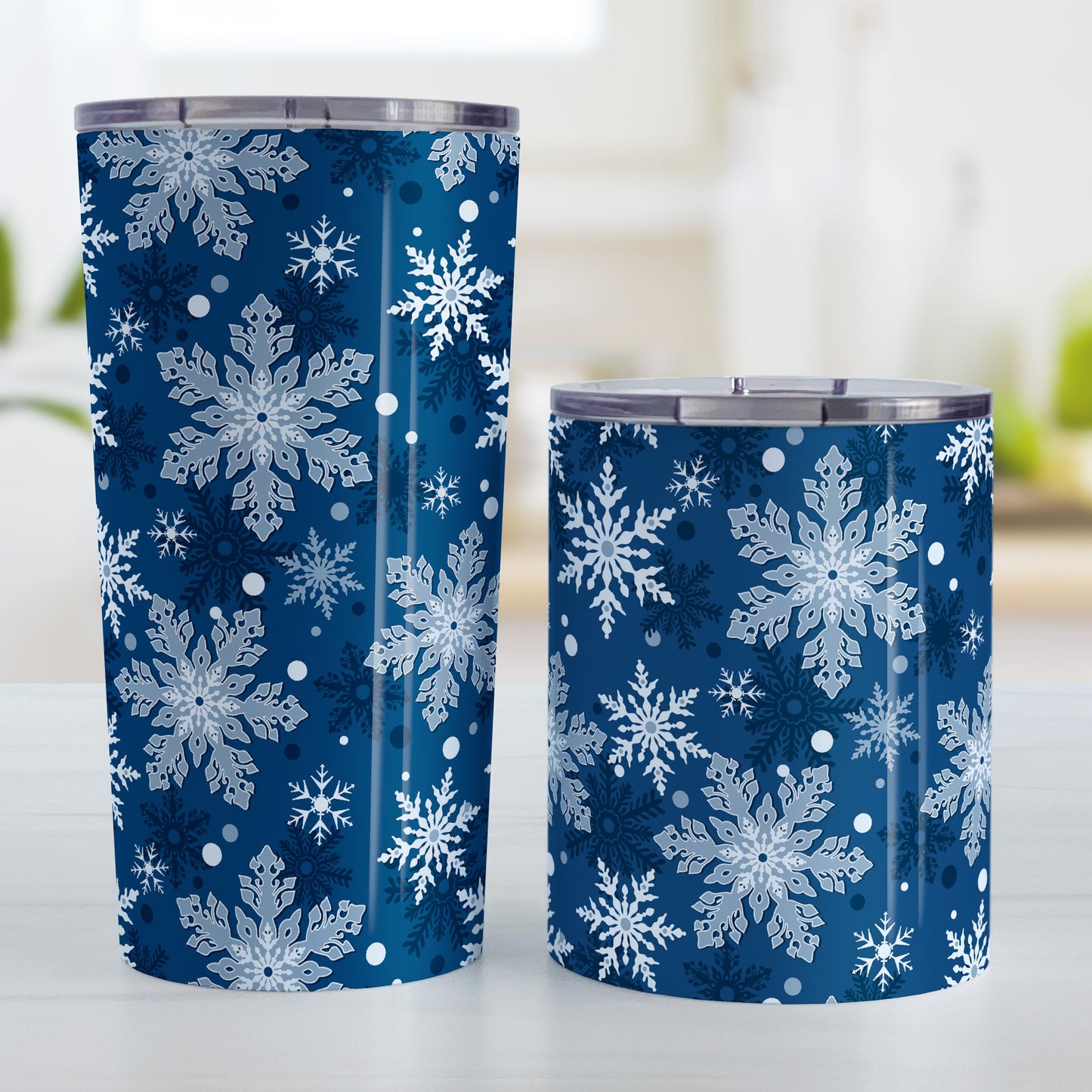 Classic Blue Snowflake Pattern Winter Tumbler Cup (20oz and 10oz, stainless steel insulated) at Amy's Coffee Mugs. A tumbler cup designed with a pattern of different shades of blue snowflakes over a classic blue background color that wraps around the cup. Photo shows both sized cups on a table next to each other.
