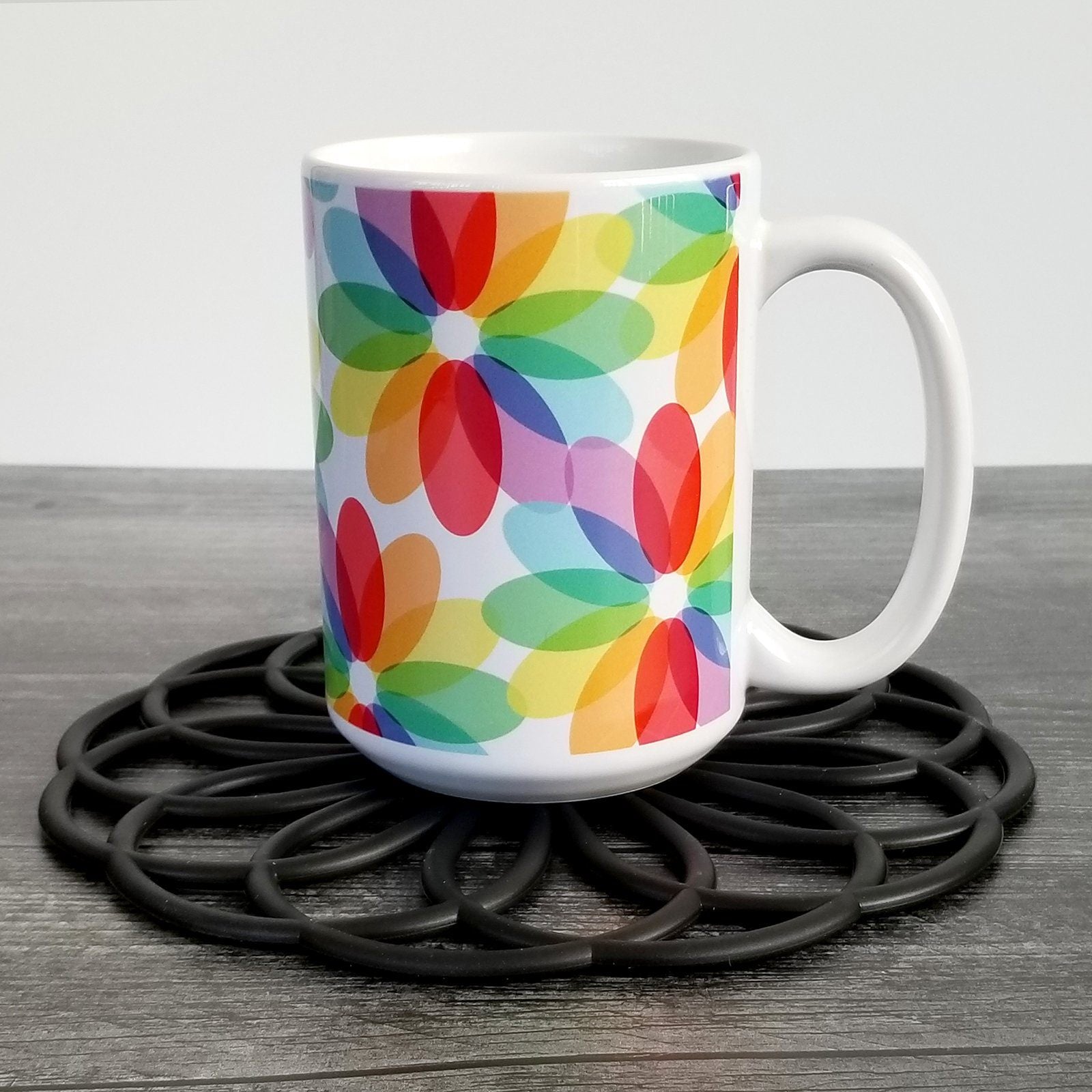 Color Lovers Rainbow Flower Mug (15oz) on weathered wood and black trivet - Amy's Coffee Mugs. A ceramic coffee mug with large colorful flower-like designs with each oval petal presenting a different progression in the rainbow, blending into each other, in a pattern that wraps around the mug up to the handle.
