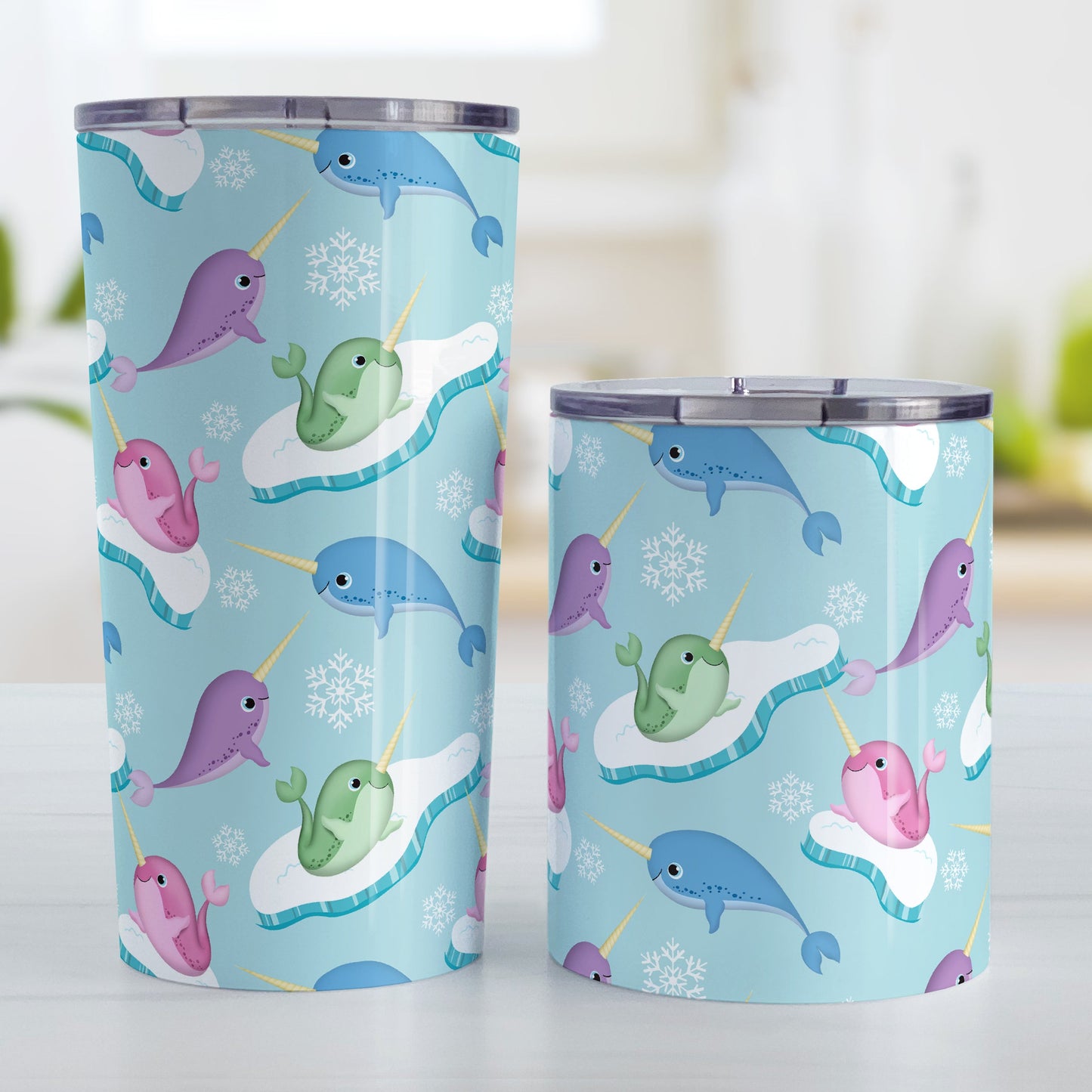 Colorful Arctic Narwhal Pattern Tumbler Cup (20oz and 10oz, stainless steel insulated) at Amy's Coffee Mugs. Tumbler cups designed cute pink, purple, blue, and green narwhals with icebergs and snowflakes over a light blue colored background that wraps around the cups. Photo shows both sized cups on a table next to each other.