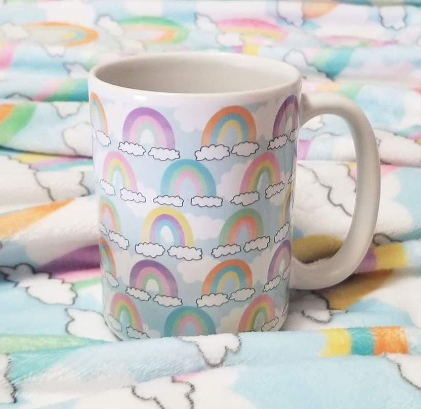 Colorful Rainbows Sky Pattern Mug at Amy's Coffee Mugs. A ceramic coffee mug designed with an illustration of different rainbows with three different semi-faded colors each sitting on clouds, over a blue sky with white clouds in the background that wraps around the mug up to the handle. Photo shows the mug sitting atop a blanket with the same illustration.