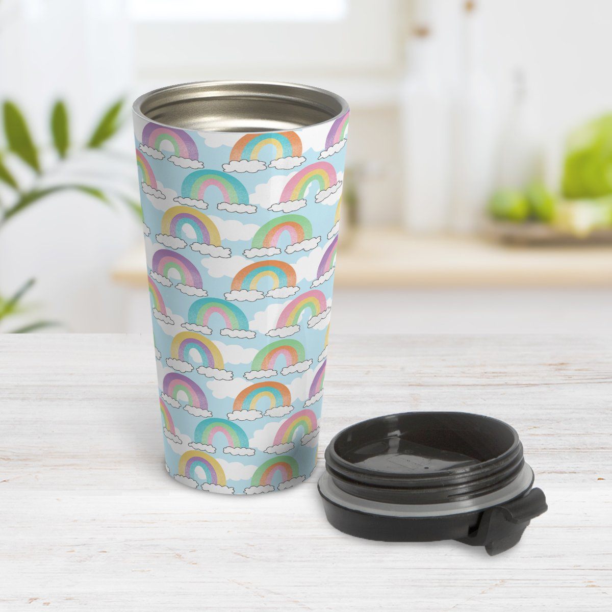 Colorful Rainbows Sky Pattern Travel Mug at Amy's Coffee Mugs. A travel mug designed with an illustration of different rainbows with three different semi-faded colors each sitting on clouds, over a blue sky with white clouds in the background that wraps around the travel mug. Photo shows the mug open on a table with the lid beside it.