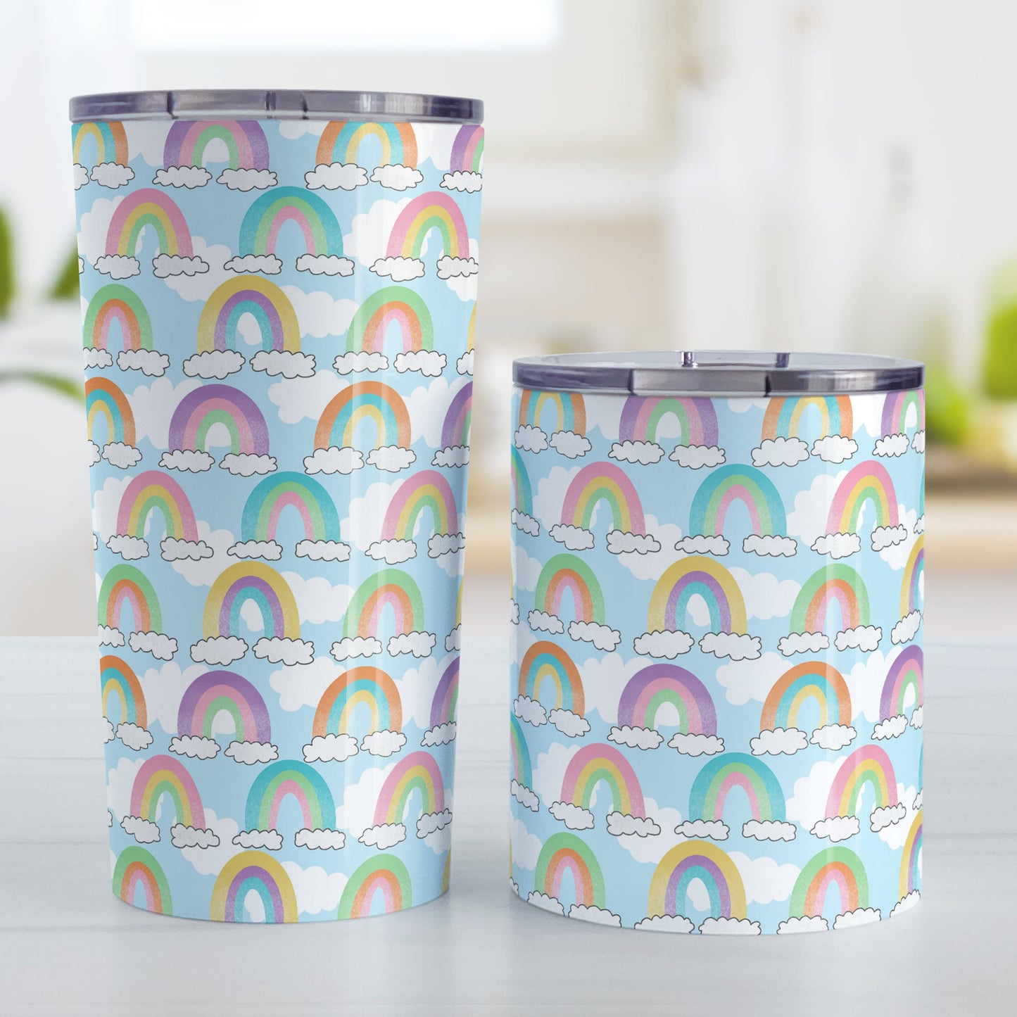 Colorful Rainbows Sky Pattern Tumbler Cup (20oz and 10oz, stainless steel insulated) at Amy's Coffee Mugs. Tumbler cups designed with an illustration of different rainbows with three different semi-faded colors each sitting on clouds, over a blue sky with white clouds in the background that wraps around the cups. Photo shows both sized cups on a table next to each other.