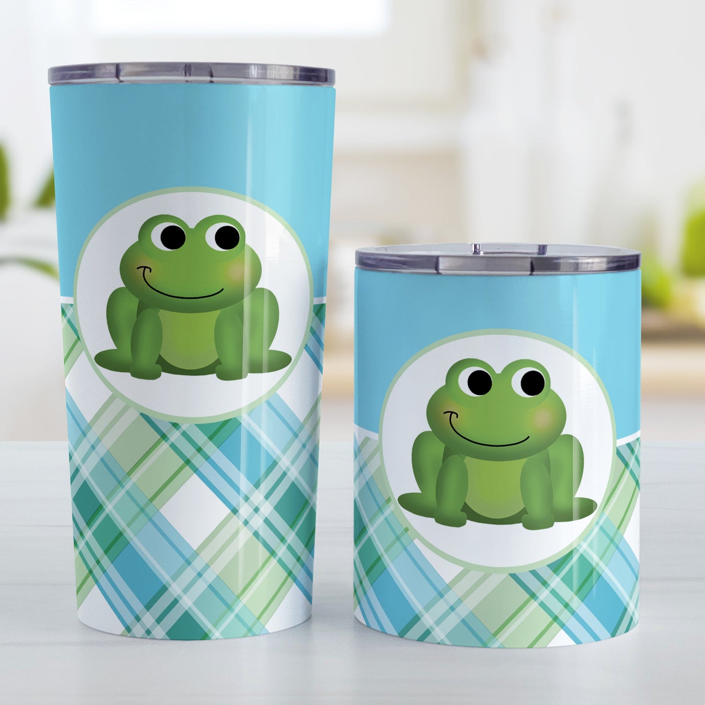 Cute Frog Green and Blue Plaid Tumbler Cup (20oz and 10oz, stainless steel insulated) at Amy's Coffee Mugs. A tumbler cup designed with a cute and smiling green frog over a background design that wraps around the cup with a solid blue color along the top and a blue and green plaid pattern along the bottom. Photo shows both sized cups on a table next to each other.