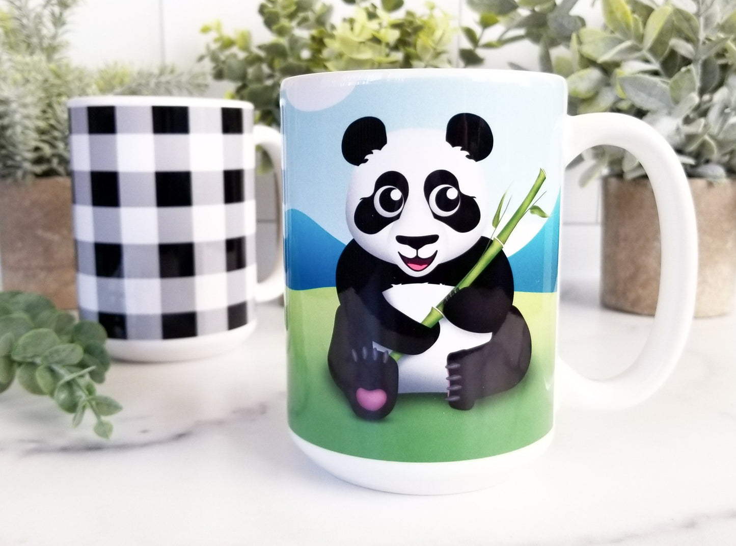Cute Panda with Bamboo - Panda Mug at Amy's Coffee Mugs. A ceramic coffee mug designed with an illustration of a panda holding bamboo sitting in the grass on both sides of the mug while the background illustration with mountains and a blue sky wraps around the mug up to the handle. Photo shows the mug on a counter with another mug and potted plants.