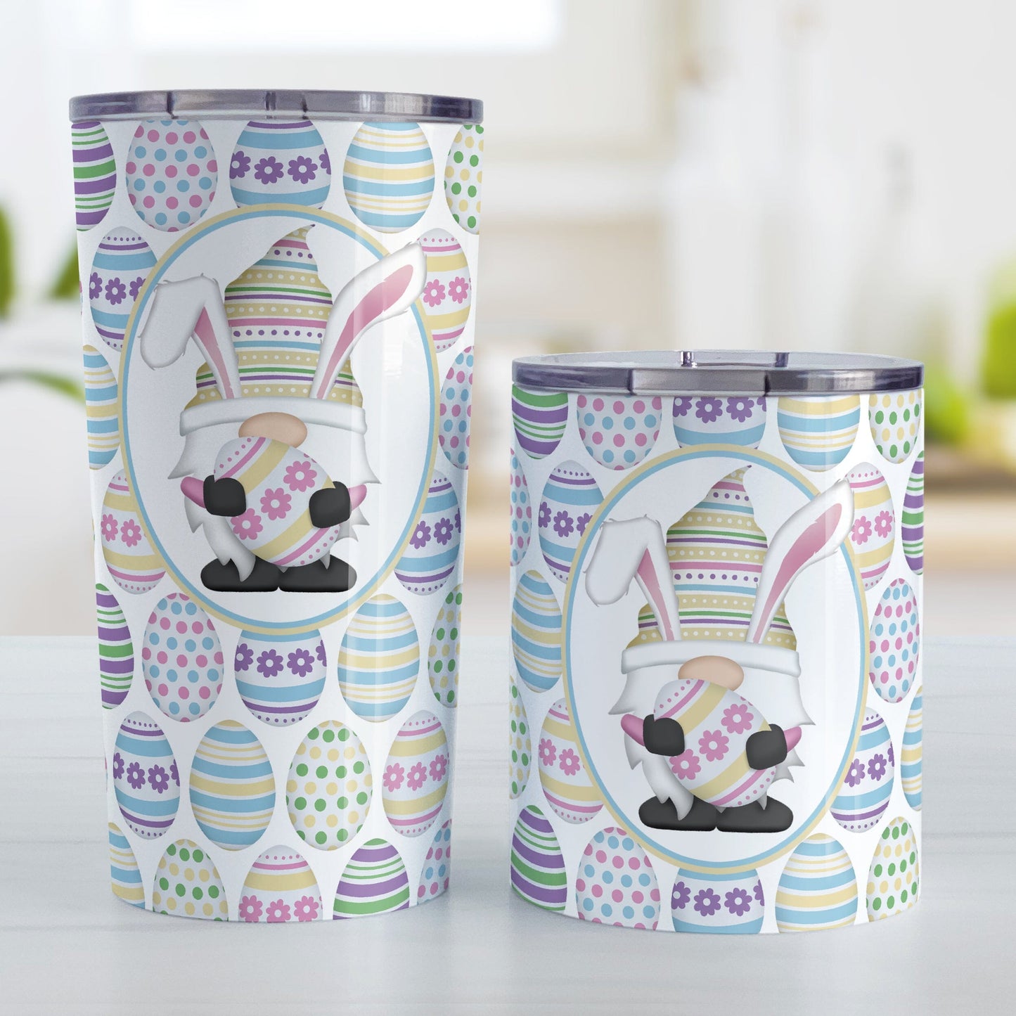 Easter Gnome Eggs Tumbler Cup (20oz or 10oz) at Amy's Coffee Mugs. Stainless steel tumbler cups designed with an illustration of an adorable gnome with bunny ears, holding a large pink and yellow Easter egg, in a white oval over a decorated Easter eggs pattern that wraps around the cups. Photo shows both sized cups next to each other on a table.