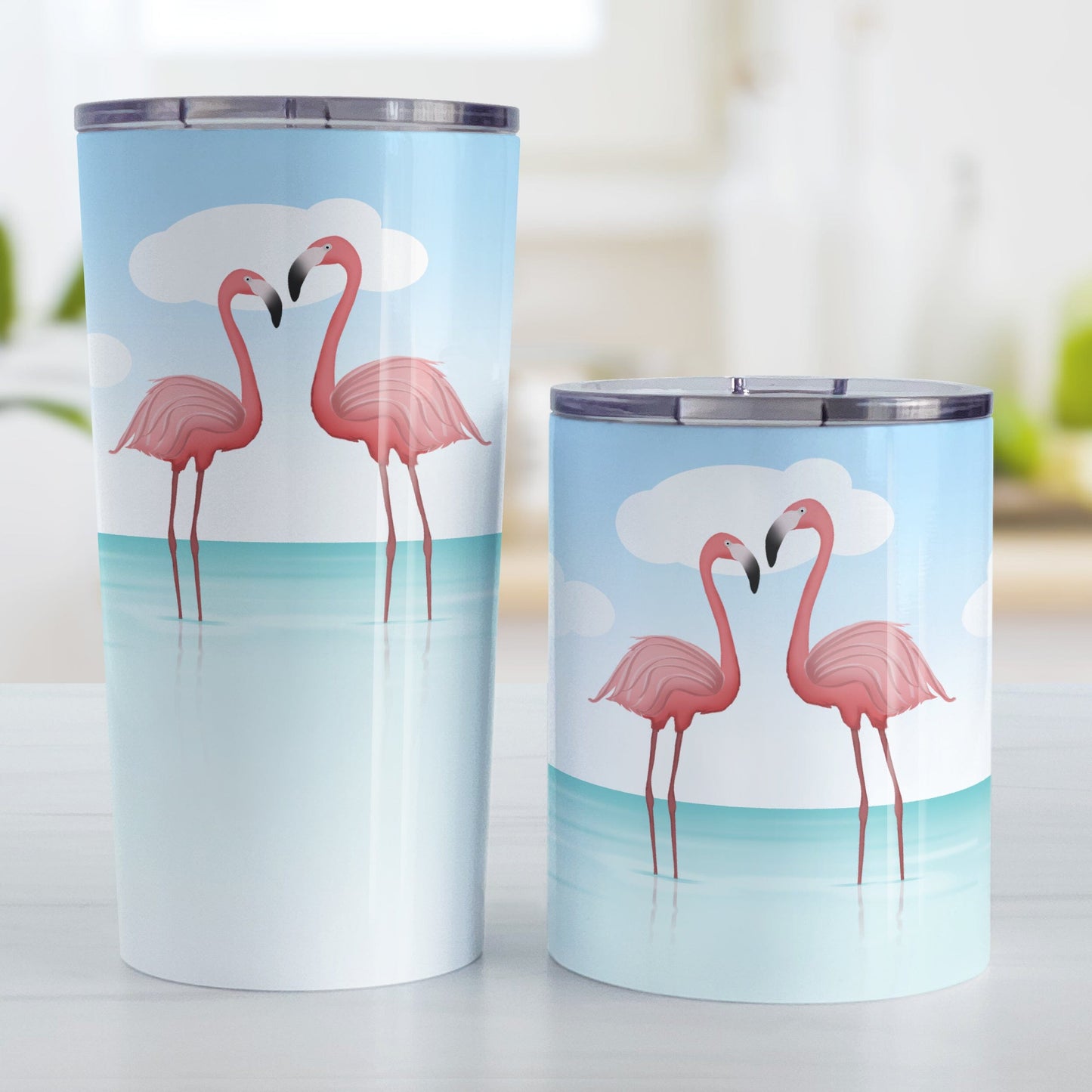 Flamingos in the Water Tumbler Cups (20oz or 10oz) at Amy's Coffee Mugs. Stainless steel tumbler cups designed with an illustration of two pink flamingos standing in the ocean water, with fresh and clean open water with ripples and a bright blue sky. Photo shows both sized cups on a table next to each other.