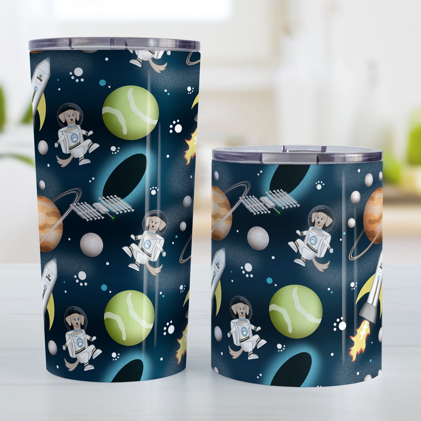 Galaxy Dog-stronaut Tumbler Cup (20oz and 10oz) at Amy's Coffee Mugs. Stainless steel insulated tumbler cups with a fun galaxy "dog-stronaut" design with dog astronauts, dog themed planets, dog bone satellites, paw print stars, black holes, and dog branded rocket ships on an outer-space background. Perfect for people who love galaxy and space designs, and love dogs too! Photo shows both sized cups next to each other.