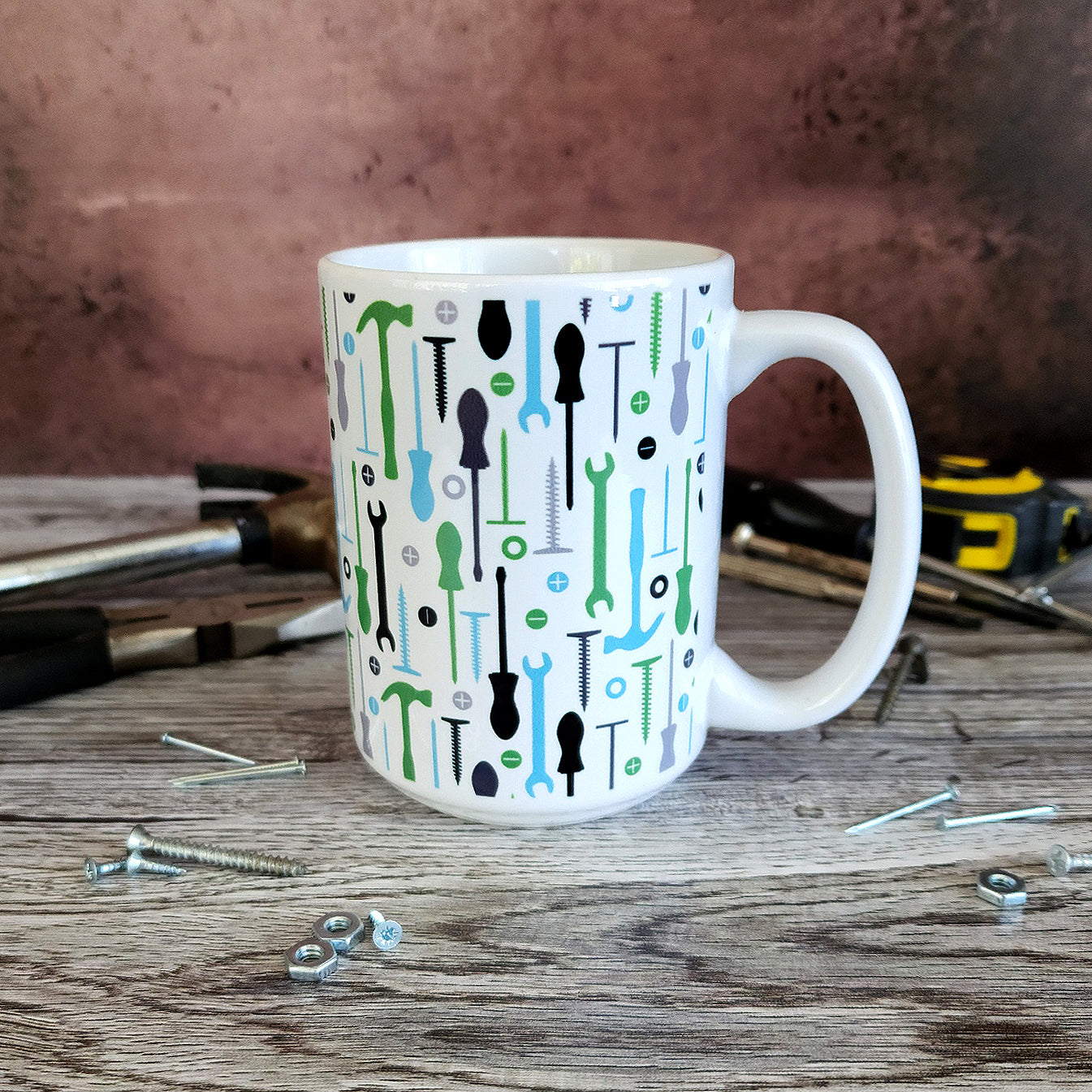 Green Blue Tools Pattern Mug (15oz on work bench with tools round it) at Amy's Coffee Mugs. A ceramic coffee mug with a modern style pattern of tools in green, blue, black, and gray over white that wraps around the mug to the handle. Perfect for any handyman or contractor.
