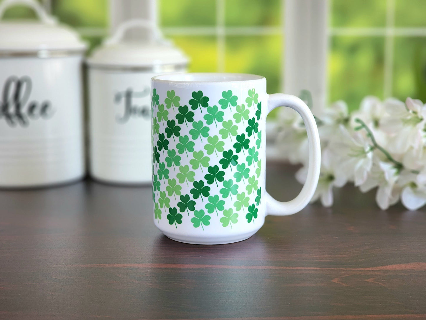 Green Clovers Mug (15oz) at Amy's Coffee Mugs. A ceramic coffee mug designed with a pattern of clovers (shamrocks) in three different shades of green that wraps around the mug up to the handle. Photo shows the mug on a table with coffee and tea jars in the background before a window.