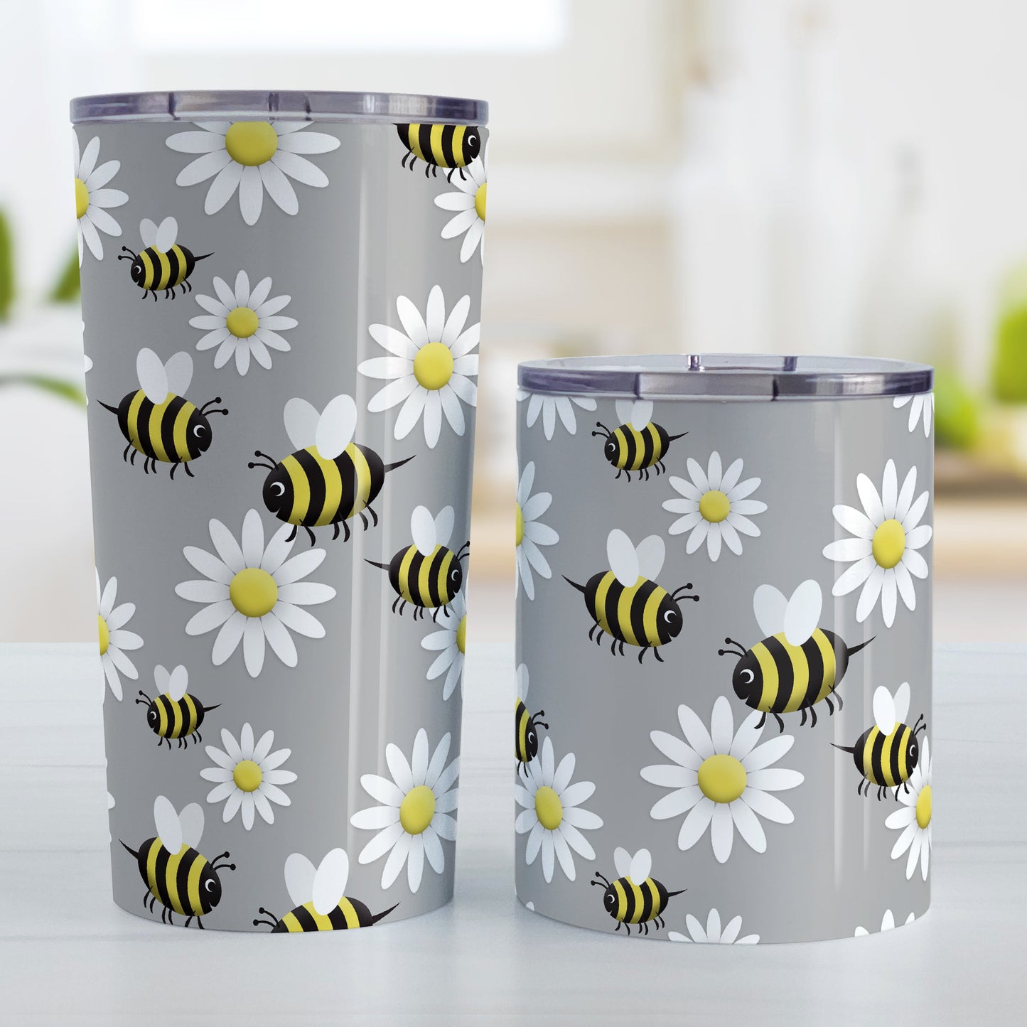 Happy Bee and Daisy Pattern Tumbler Cup (20oz and 10oz, stainless steel insulated) at Amy's Coffee Mugs. A tumbler cup designed with a cute pattern of happy bees and white daily flowers over a light gray background color that wraps around the cup. Photo shows both sized cups on a table next to each other.