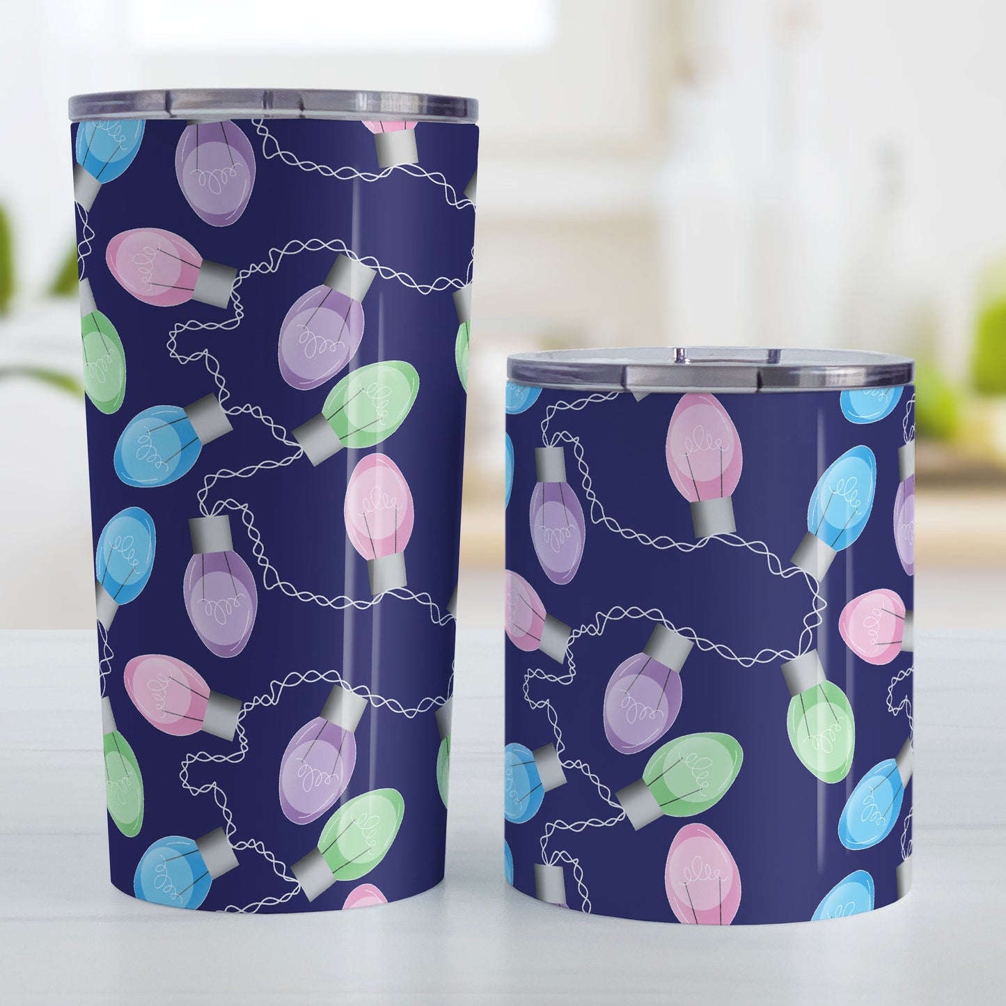 Holiday Lights Pattern Christmas Tumbler Cup (20oz and 10oz, stainless steel insulated) at Amy's Coffee Mugs. A tumbler cup designed with string of colorful holiday lights in pink, blue, green, and purple in a pattern over a navy blue background color that wraps around the mug up to the cup. Photo shows both sized cups on a table next to each other.