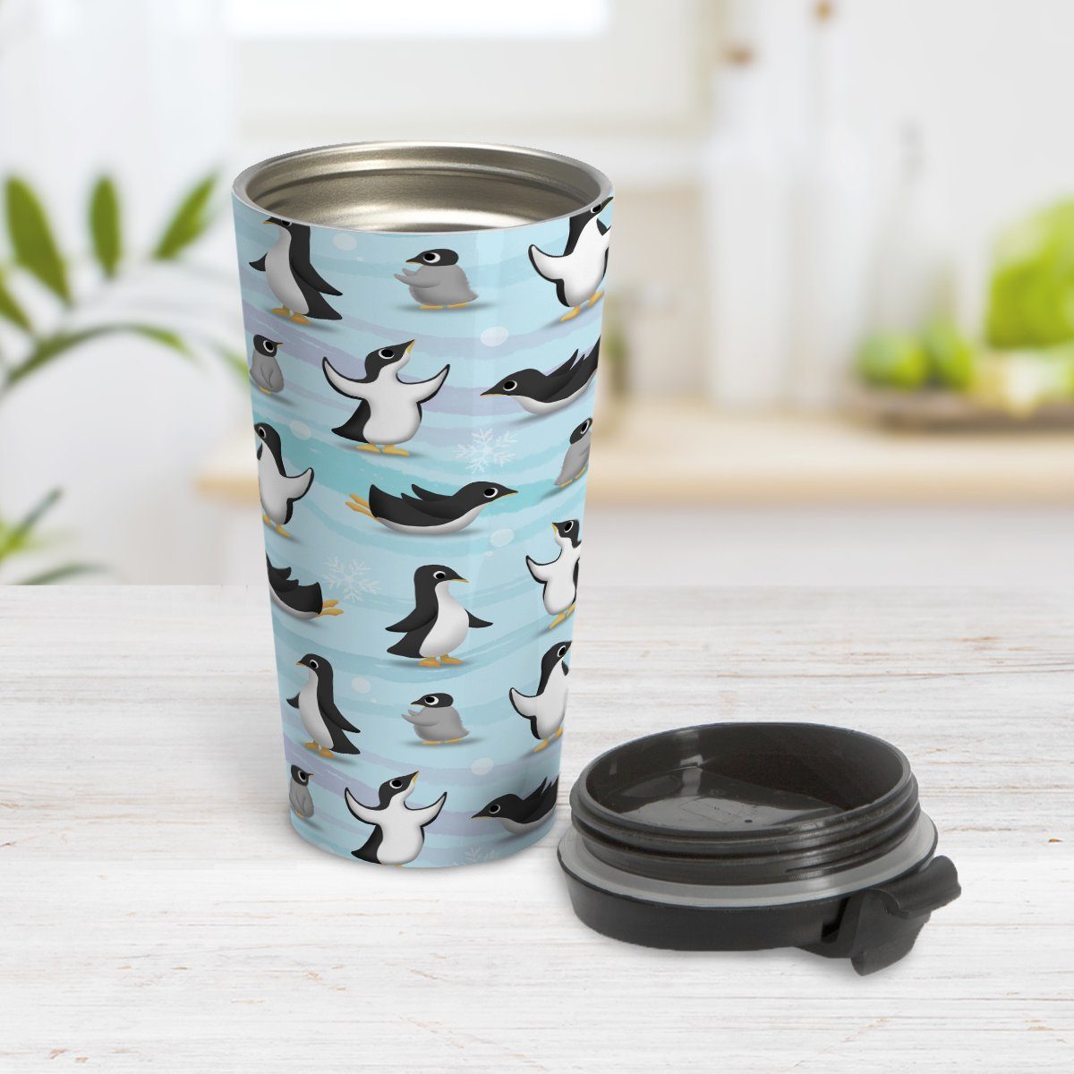 Penguin Parade Pattern Travel Mug at Amy's Coffee Mugs. A travel mug designed with a fun penguin parade pattern featuring a variety of penguins and baby penguins over an Antarctic background with waves of blue, turquoise, and purple colors with hints of snowflakes that wraps around the mug. Photo shows the mug open on a table with its lid beside it.