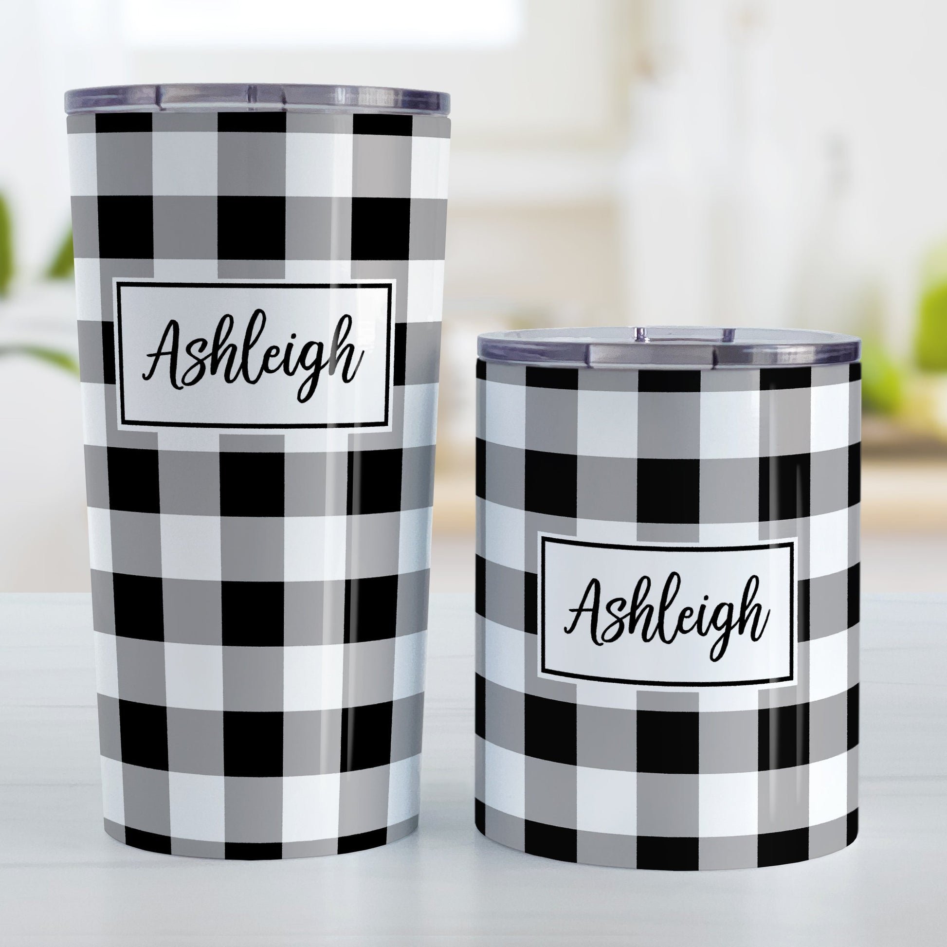 Personalized Black and White Buffalo Plaid Tumbler Cup (20oz and 10oz, stainless steel insulated) at Amy's Coffee Mugs. A tumbler cup designed with a black and white buffalo plaid (buffalo check) pattern that wraps around the cup. Your name is printed in a black script font in a white frame design over the buffalo plaid background. Photo shows both sized cups on a table next to each other.