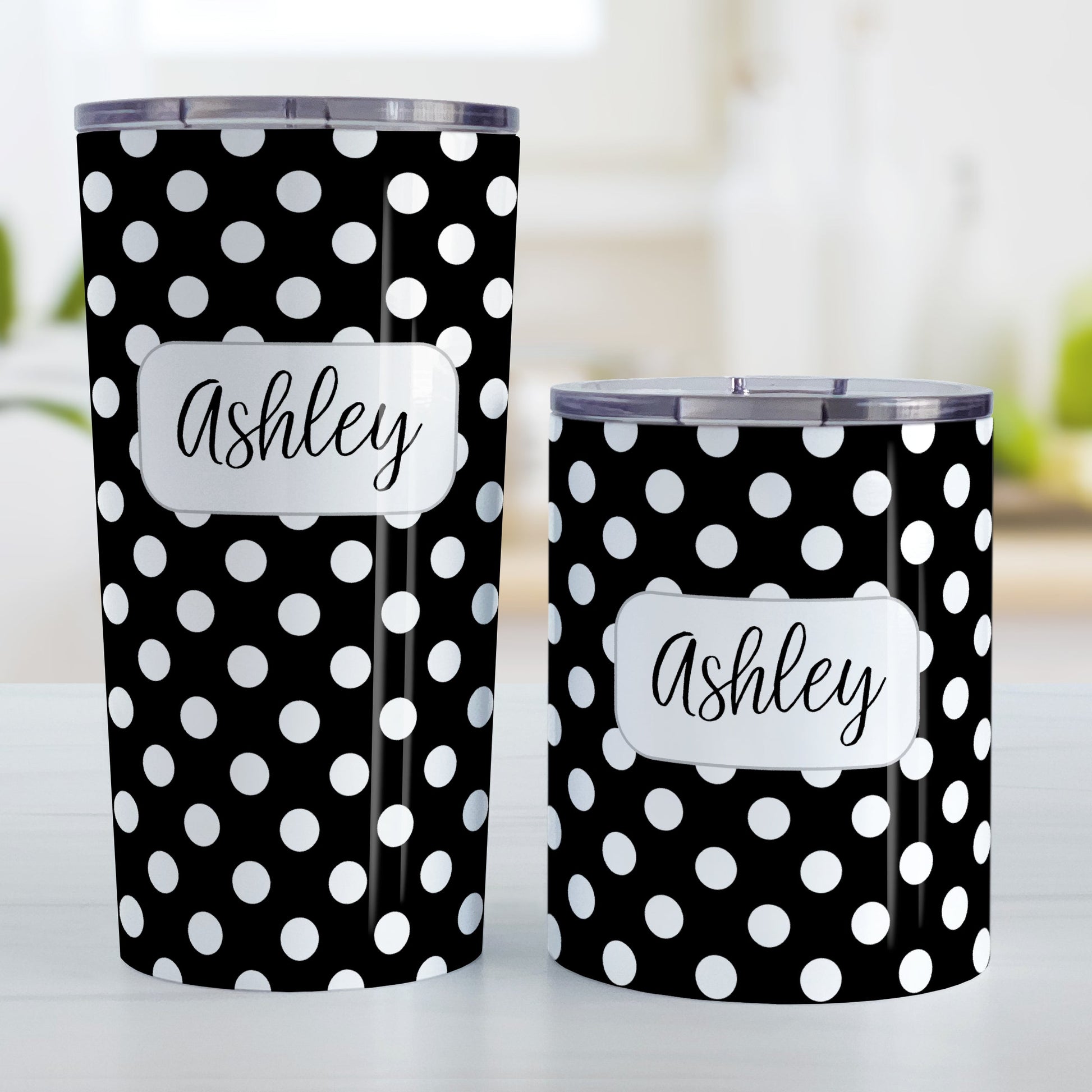 Personalized Black Polka Dot Tumbler Cup (20oz and 10oz, stainless steel insulated) at Amy's Coffee Mugs. A tumbler cup designed with a pattern of white polka dots over a black background color that wraps around the cup. Your name is personalized in a cute black script font in a white frame design over the polka dot pattern. Photo shows both sized cups on a table next to each other.