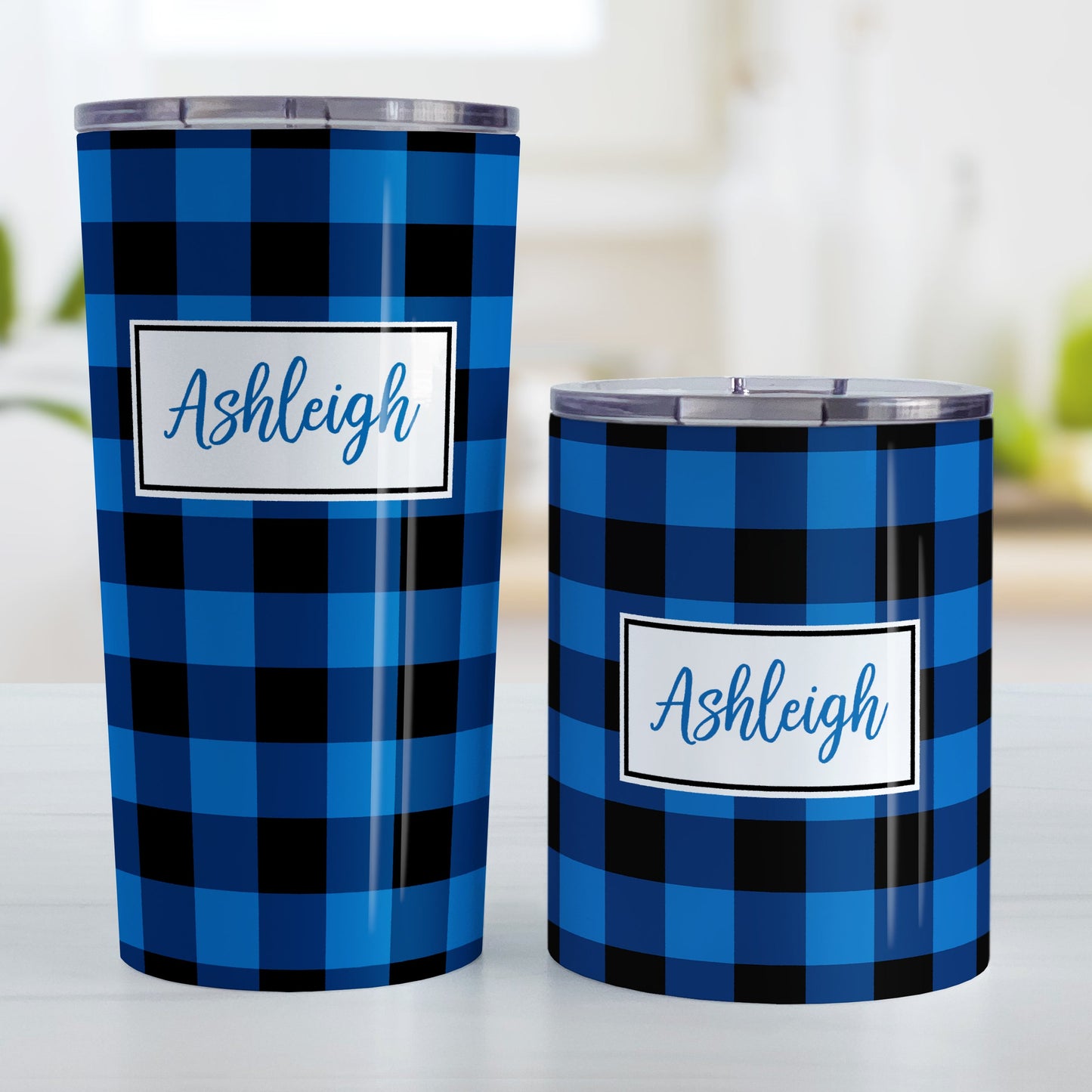 Personalized Blue and Black Buffalo Plaid Tumbler Cup (20oz and 10oz, stainless steel insulated) at Amy's Coffee Mugs. A tumbler cup designed with a blue and black buffalo plaid (buffalo check) pattern that wraps around the cup. Your name is printed in a blue script font in a white frame design over the buffalo plaid background. Photo shows both sized cups on a table next to each other.