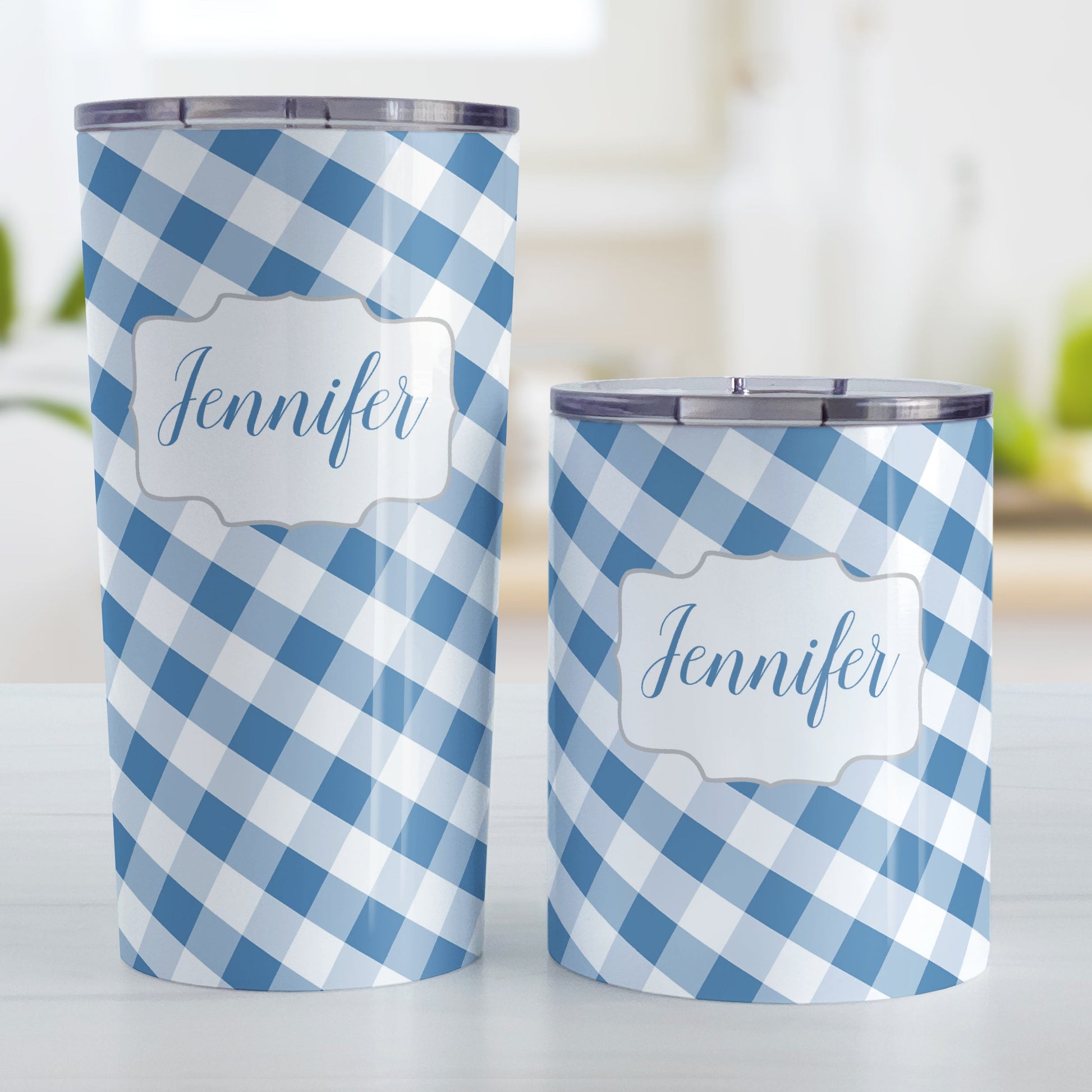 Personalized Blue Gingham Tumbler Cup (20oz and 10oz, stainless steel insulated) at Amy's Coffee Mugs. A tumbler cup designed with a slanted blue and white gingham pattern that wraps around the cup. Your personalized name is custom-printed in a blue script font in a white frame design over the gingham pattern. Photo shows both sized cups on a table next to each other.