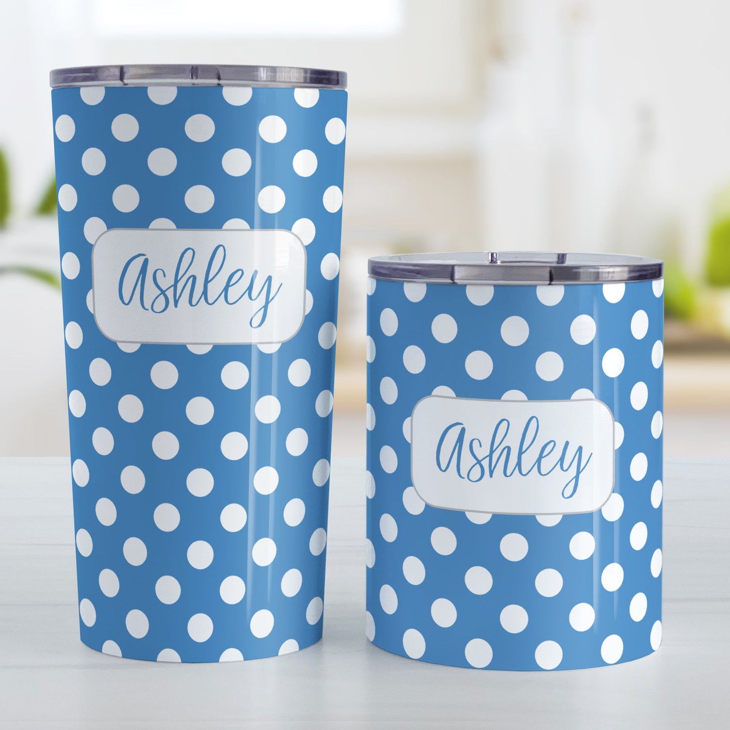 Personalized Blue Polka Dot Tumbler Cup (20oz and 10oz, stainless steel insulated) at Amy's Coffee Mugs. These cups are designed with a pattern of white polka dots over a blue background color that wraps around the cups. Your name is personalized in a blue script font over the polka dot pattern. Photo shows both sized cups on a table next to each other.