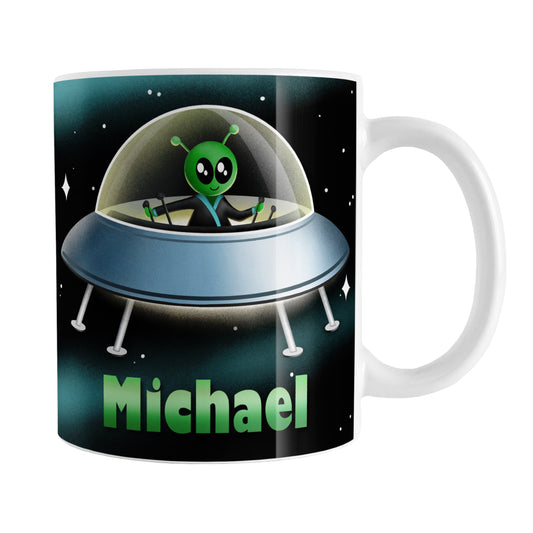 Personalized Green Alien Spaceship Mug (11oz) at Amy's Coffee Mugs. A ceramic coffee mug designed with a unique and cute green alien piloting a blue saucer spaceship with a glass dome, on both sides of the mug, over an outer space background that wraps around the mug up to the handle. Your personalized name is custom-printed in green below the alien spaceship design.