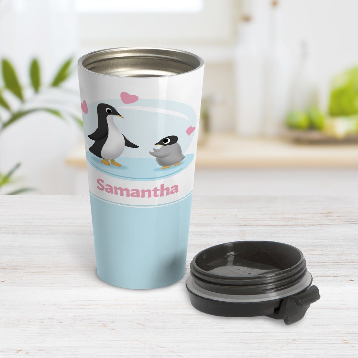 Personalized My Little Penguin Travel Mug at Amy's Coffee Mugs. A travel mug designed with an illustration of a mommy or daddy penguin with a little baby penguin over a blue background design with three pink hearts above them. Your name is personalized in pink below the adorable penguins. Photo shows the mug open on a table with its lid beside it.
