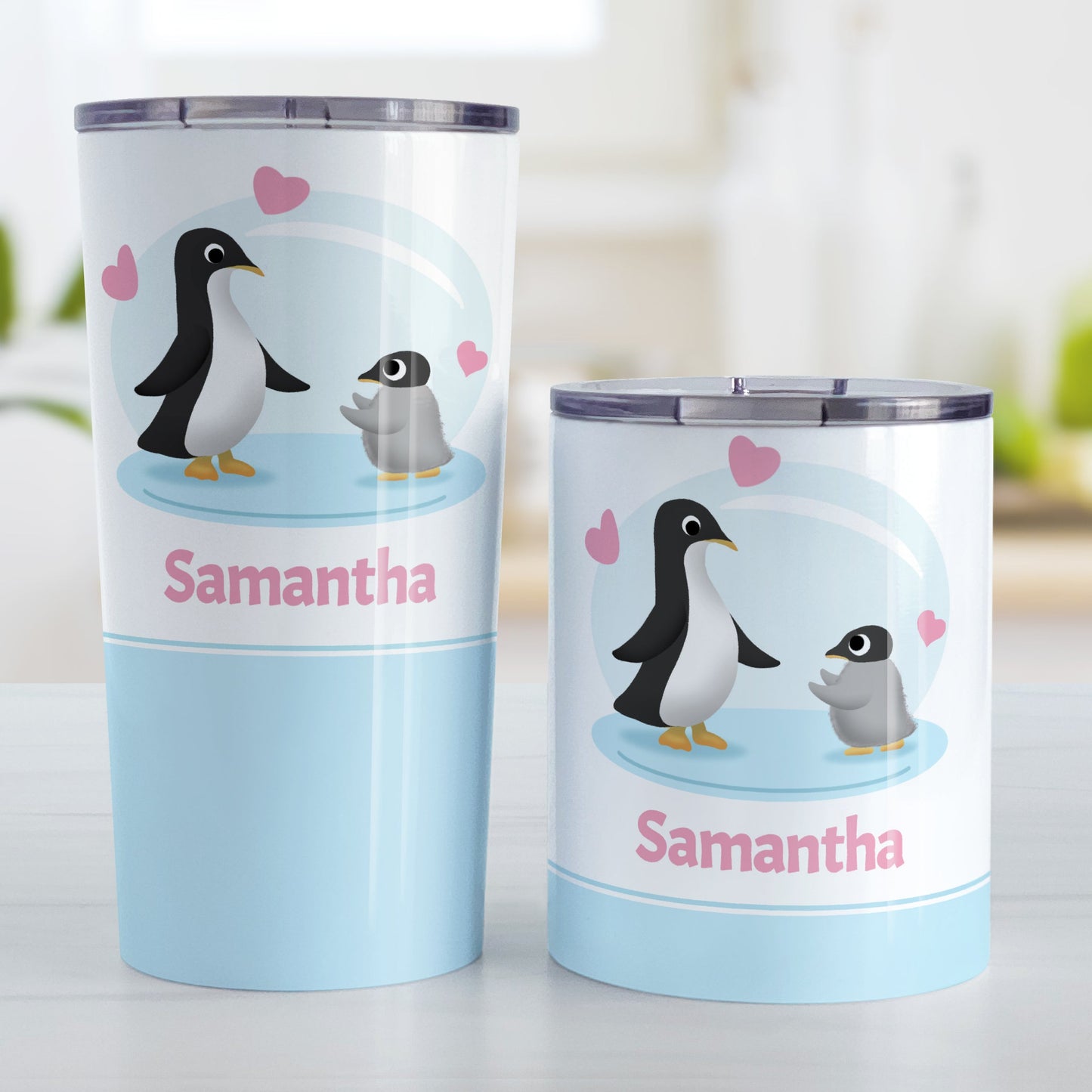 Personalized My Little Penguin Tumbler Cup (20oz and 10oz, stainless steel insulated) at Amy's Coffee Mugs. A tumbler cup designed with an illustration of a mommy or daddy penguin with a little baby penguin over a blue background design with three pink hearts above them. Your name is personalized in pink below the adorable penguins. Photo shows both sized cups on a table next to each other.