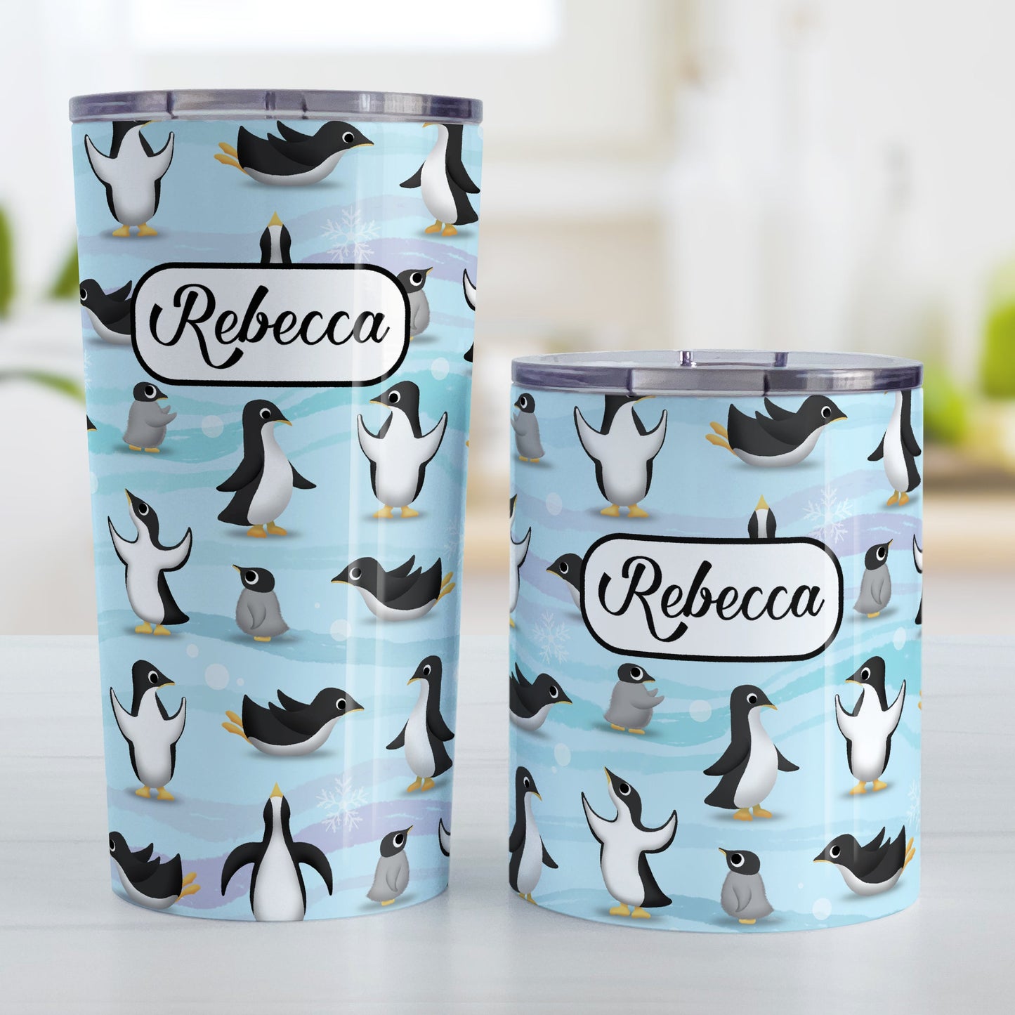 Personalized Penguin Parade Pattern Tumbler Cup (20oz and 10oz, stainless steel insulated) at Amy's Coffee Mugs. A tumbler cup designed with a fun penguin parade pattern featuring a variety of penguins and baby penguins over an Antarctic background with waves of blue accented with turquoise and purple colors with hints of snowflakes that wraps around the cup. Your name is printed in black within a white frame design over the penguin pattern. Photo shows both sized cups on a table next to each other.
