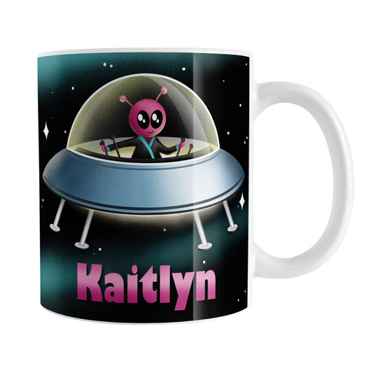 Personalized Pink Alien Spaceship Mug (11oz) at Amy's Coffee Mugs. A ceramic coffee mug designed with a unique and cute pink alien piloting a blue saucer spaceship with a glass dome, on both sides of the mug, over an outer space background that wraps around the mug up to the handle. Your personalized name is custom-printed in pink below the alien spaceship design.