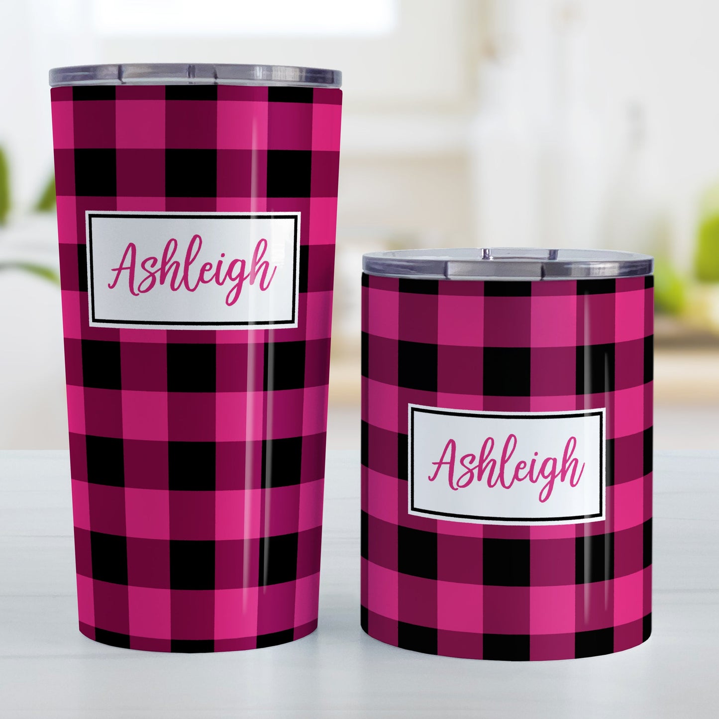 Personalized Pink and Black Buffalo Plaid Tumbler Cup (20oz and 10oz, stainless steel insulated) at Amy's Coffee Mugs. A tumbler cup designed with a raspberry pink and black buffalo plaid (buffalo check) pattern that wraps around the cup. Your name is printed in a raspberry pink script font in a white frame design over the buffalo plaid background. Photo shows both sized cups on a table next to each other.