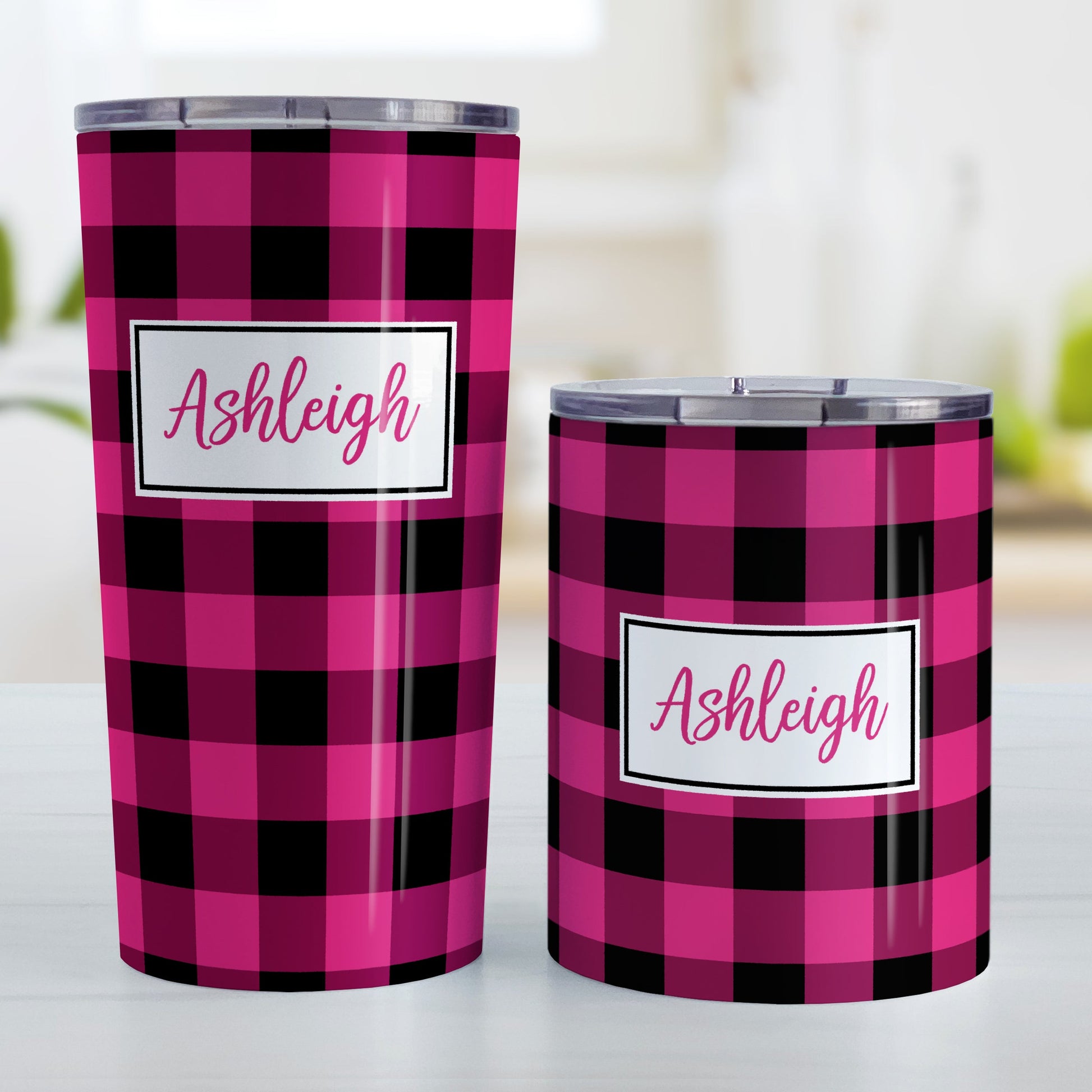 Personalized Pink and Black Buffalo Plaid Tumbler Cup (20oz and 10oz, stainless steel insulated) at Amy's Coffee Mugs. A tumbler cup designed with a raspberry pink and black buffalo plaid (buffalo check) pattern that wraps around the cup. Your name is printed in a raspberry pink script font in a white frame design over the buffalo plaid background. Photo shows both sized cups on a table next to each other.