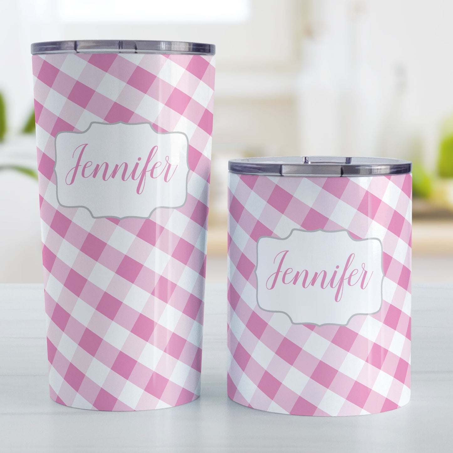 Personalized Pink Gingham Tumbler Cup (20oz and 10oz, stainless steel insulated) at Amy's Coffee Mugs. A tumbler cup designed with a slanted pink and white gingham pattern that wraps around the cup. Your personalized name is custom-printed in a pink script font in a white frame design over the gingham pattern. Photo shows both sized mugs on a table next to each other.
