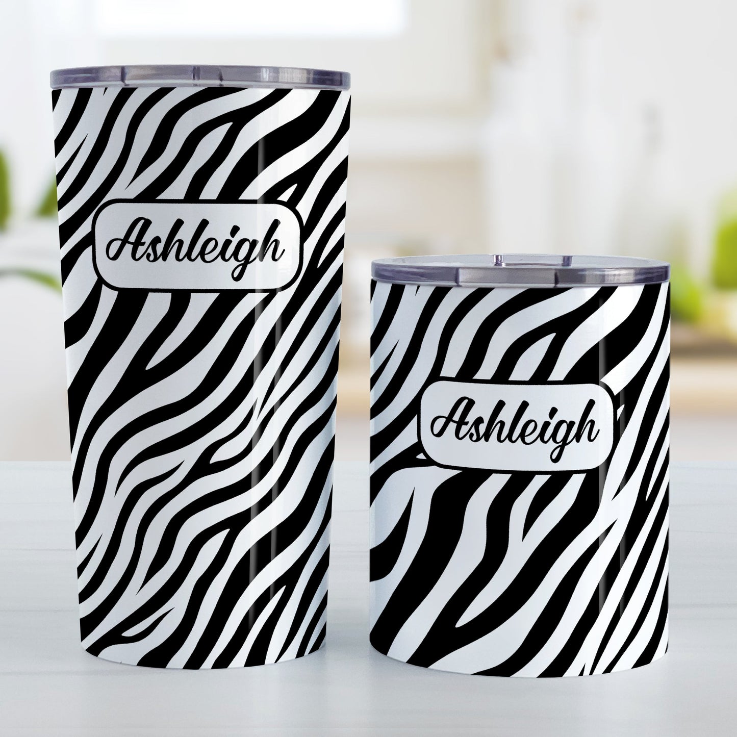 Personalized Zebra Print Pattern Tumbler Cup (20oz and 10oz, stainless steel insulated) at Amy's Coffee Mugs. A tumbler cup designed with an animal print pattern of black and white zebra stripes that wraps around the cup. Your name is custom-printed in a stylish black script font in a white rectangular design over the zebra print pattern. Photo shows both sized cups on a table next to each other.