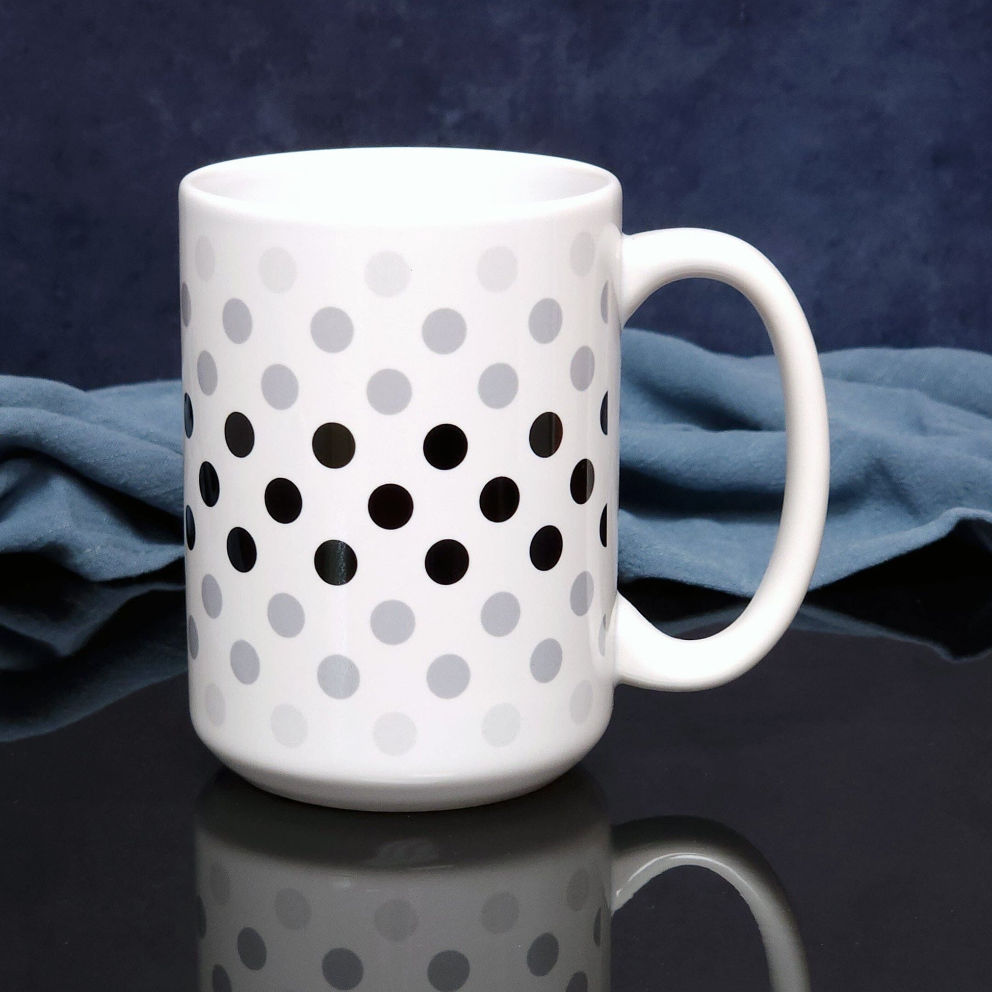 Polka Dots in Black Gray Mug (15oz) on a glossy black tabletop with a midnight blue background.. A ceramic coffee mug designed with polka dots in different shades of black and gray, with the black across the middle and the lighter shades of gray along the top and bottom, in a pattern that wraps around the mug to the handle.
