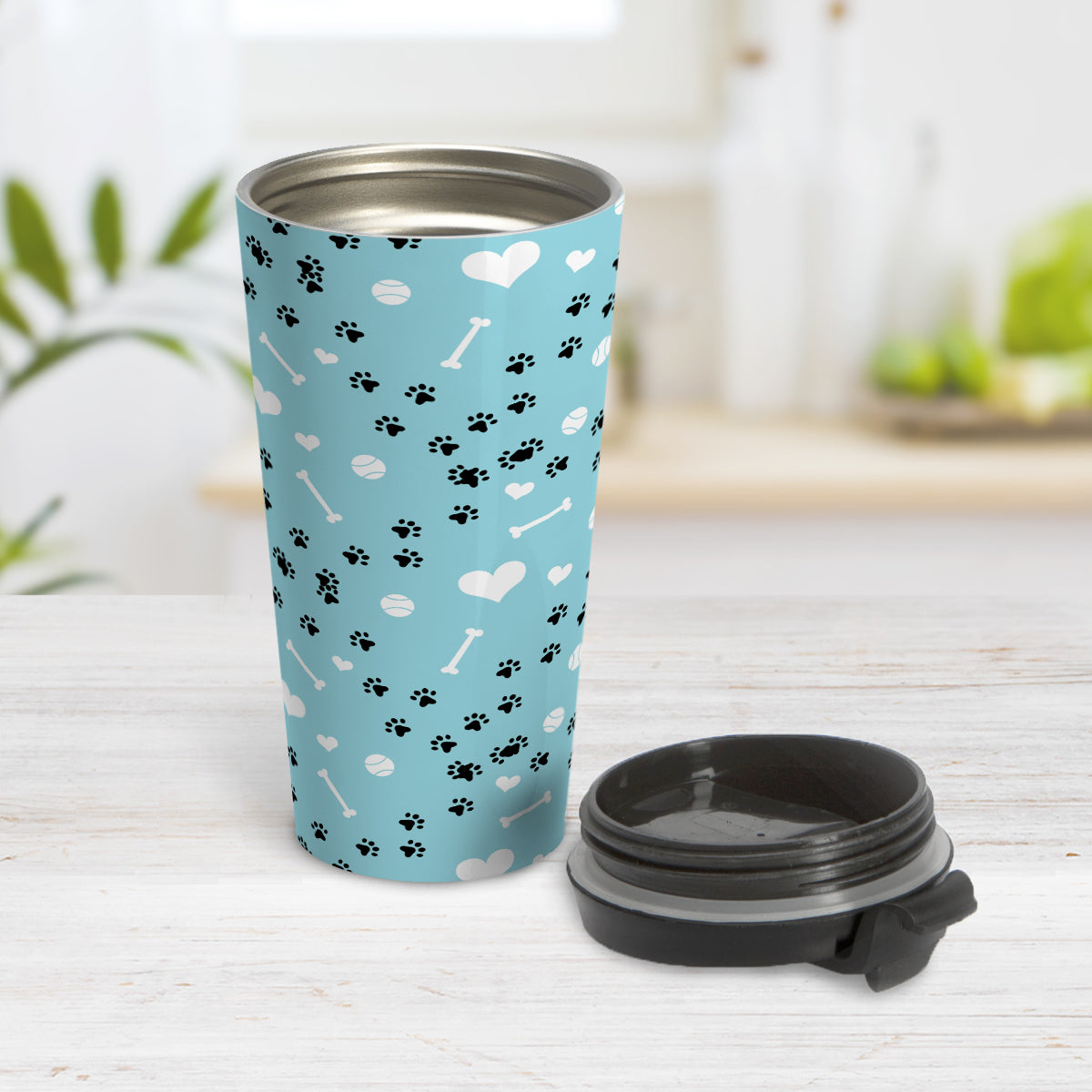 Puppy Run Blue Dog Travel Mug (15oz) at Amy's Coffee Mugs. A stainless steel travel mug designed with a pattern of chaotic tracks of puppy paw prints running around the mug with white dog bones, balls, and hearts over a light blue background. This mug is perfect for people who love the chaotic play of puppies, love dogs, and like blue travel mugs. Photo shows open travel mug with the lid on the table beside it.